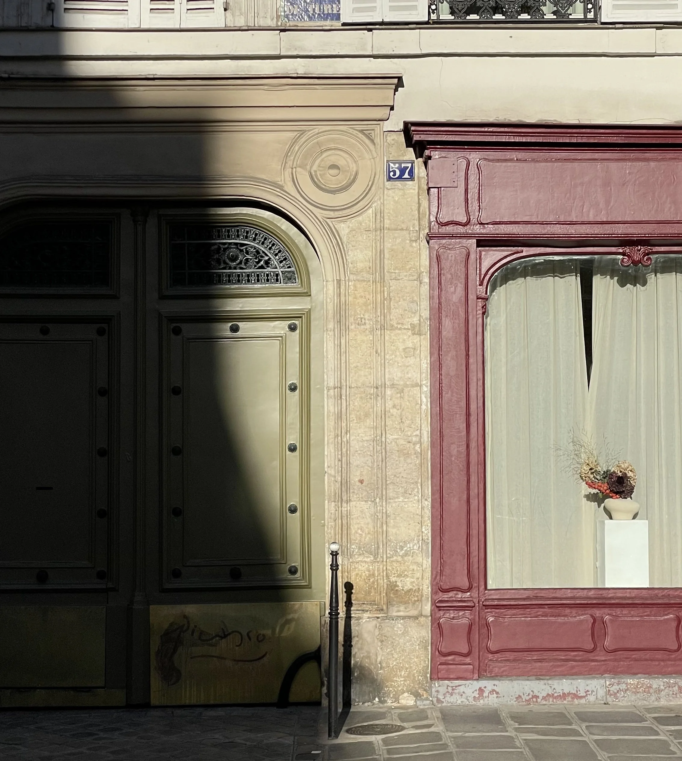 The image depicts a shop front with contrasting colors and architectural details. The left side features a dark-colored door with rounded and rectangular paneling, situated within an ornate archway with intricate decorative molding above. The right side showcases a red-painted storefront with large glass windows and elegant trim. In front, there is a sidewalk with tiled stones, a black pole, a small chain, and a porch with a basket of flowers, indicating an inviting entrance.