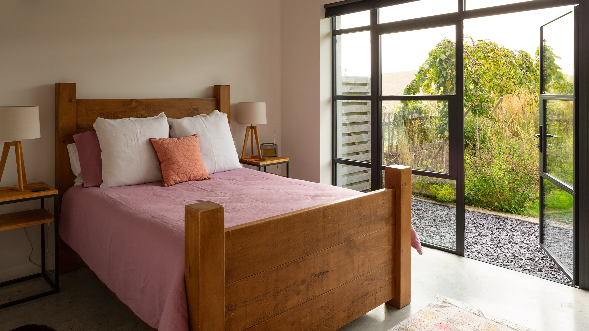 Cozy bedroom with wooden bed, pink bedding, white pillows, bedside tables, lamps, and a large window overlooking a garden.