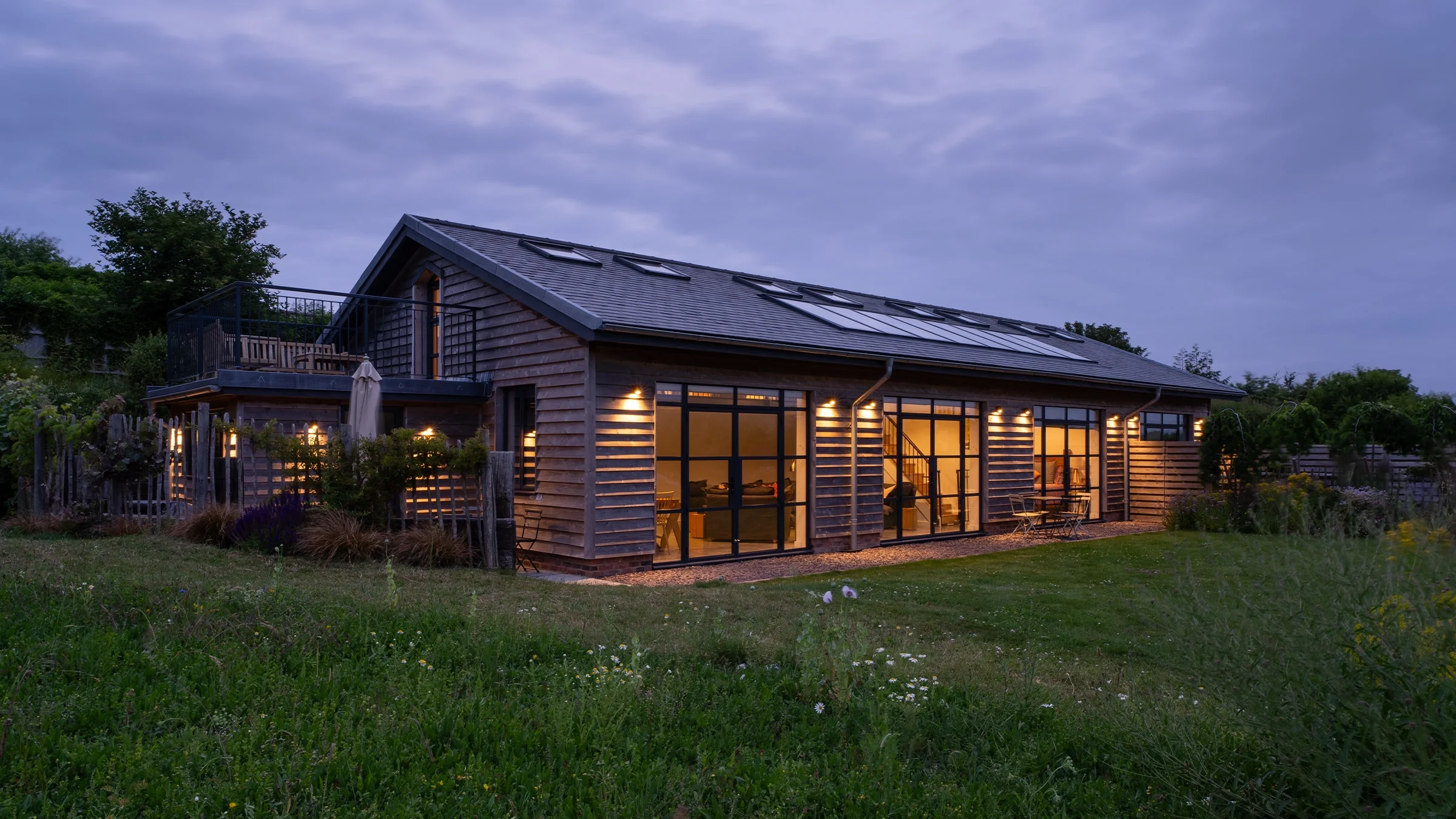 A modern wooden house with large glass windows and a rooftop deck, illuminated from within, set in a lush green yard during dusk with a cloudy sky overhead.