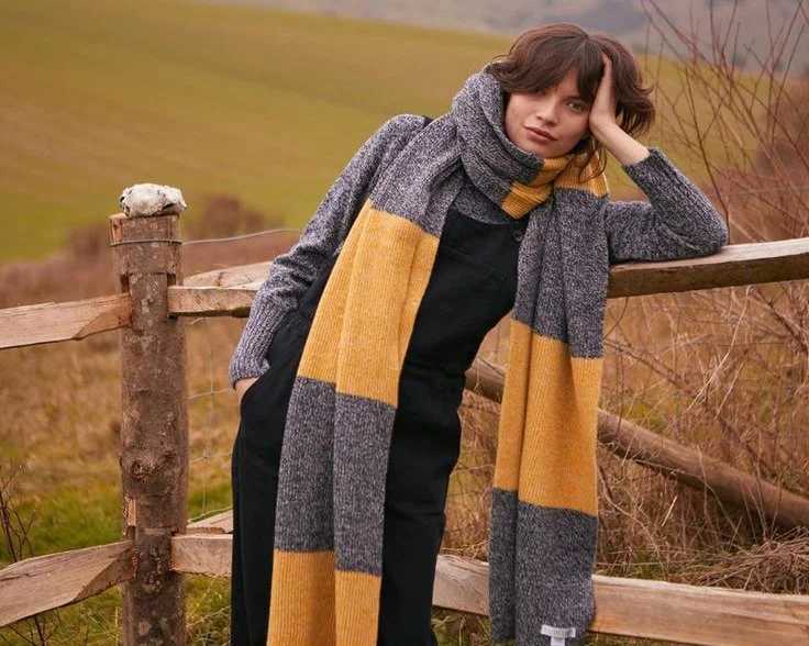 Person wearing a long gray and yellow scarf, leaning on a wooden fence in a field