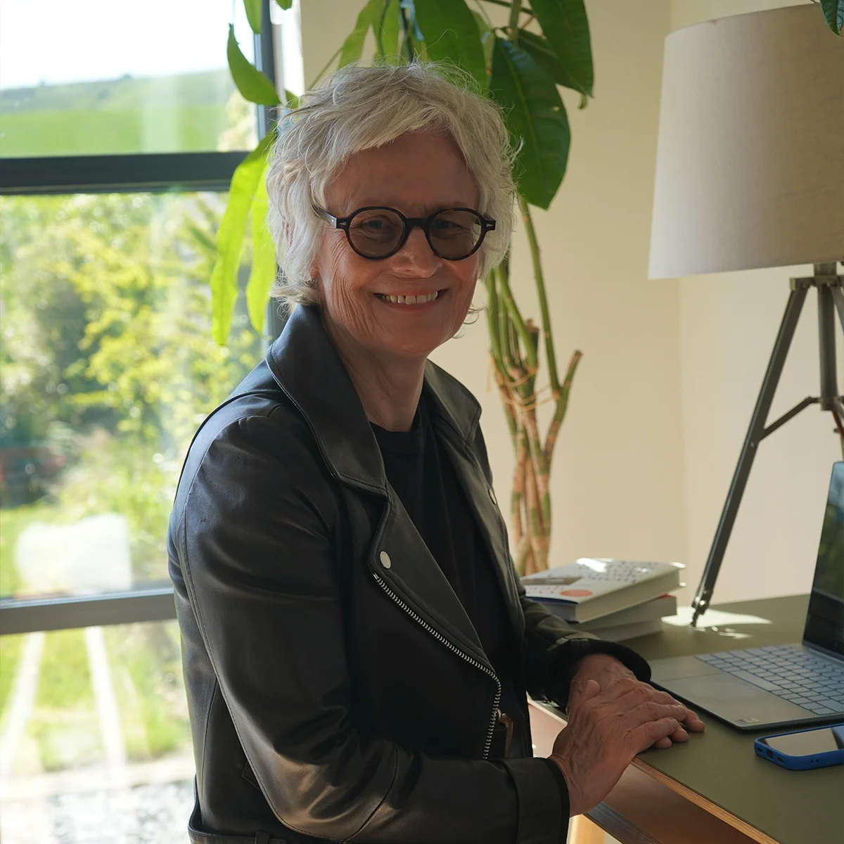 An elderly woman with gray hair and glasses smiling while sitting at a desk, with a laptop and a stack of books, near a large window with green trees outside.