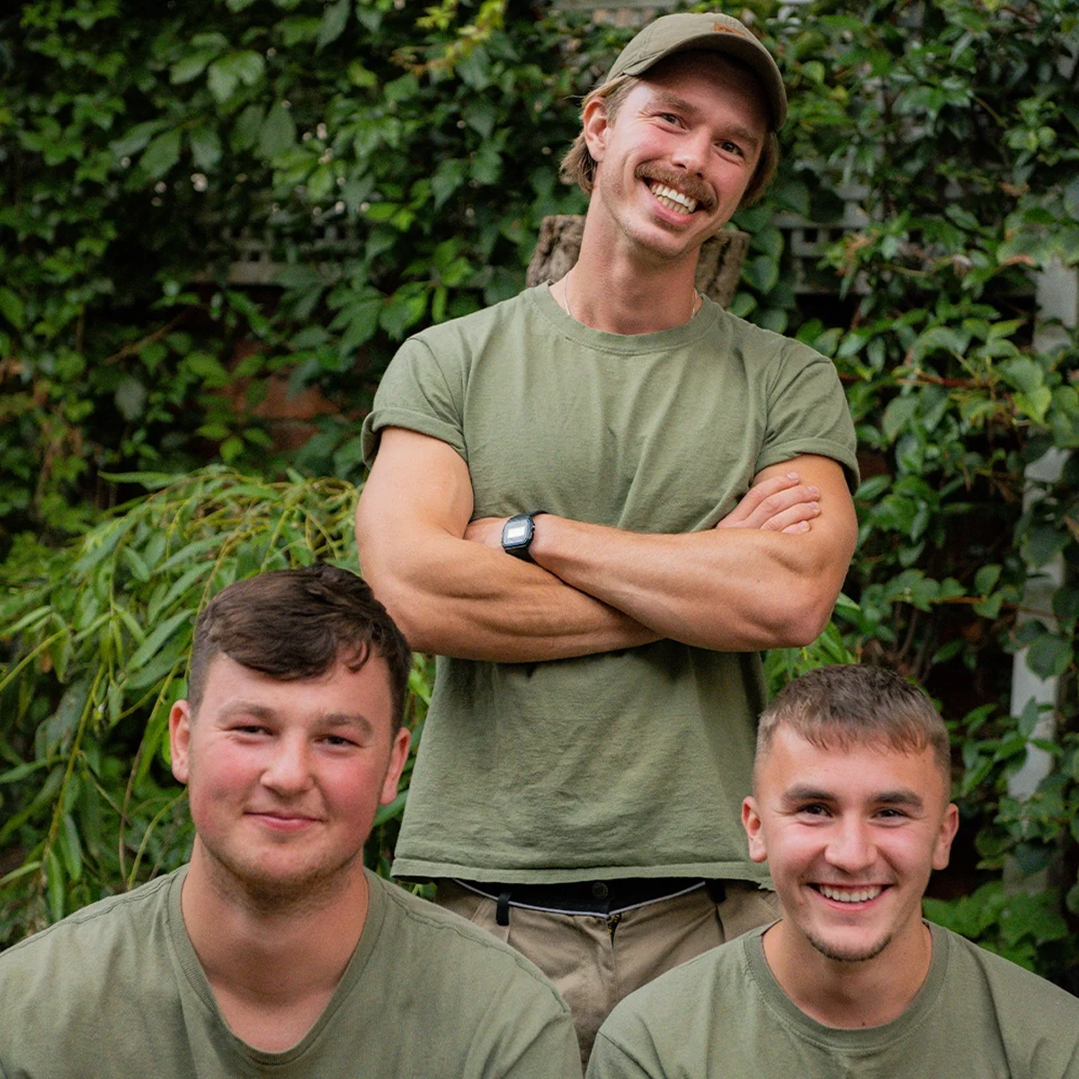 Three young men outdoors in front of green foliage; one man stands in the back smiling, with arms crossed, wearing a cap and a watch, while the two men sit in front, smiling at the camera.