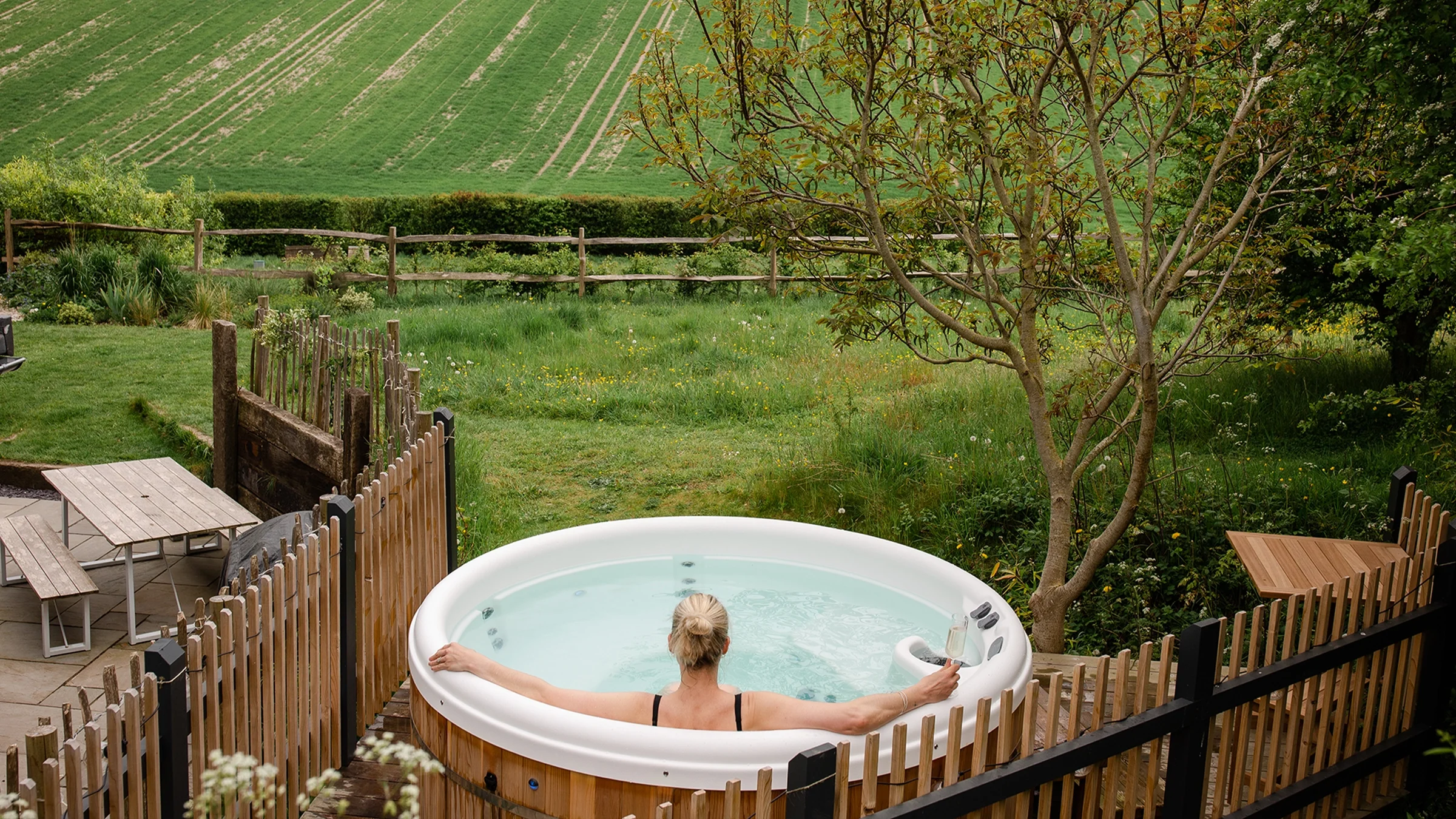 Person relaxing in an outdoor hot tub overlooking a grassy field, surrounded by a wooden fence and trees.