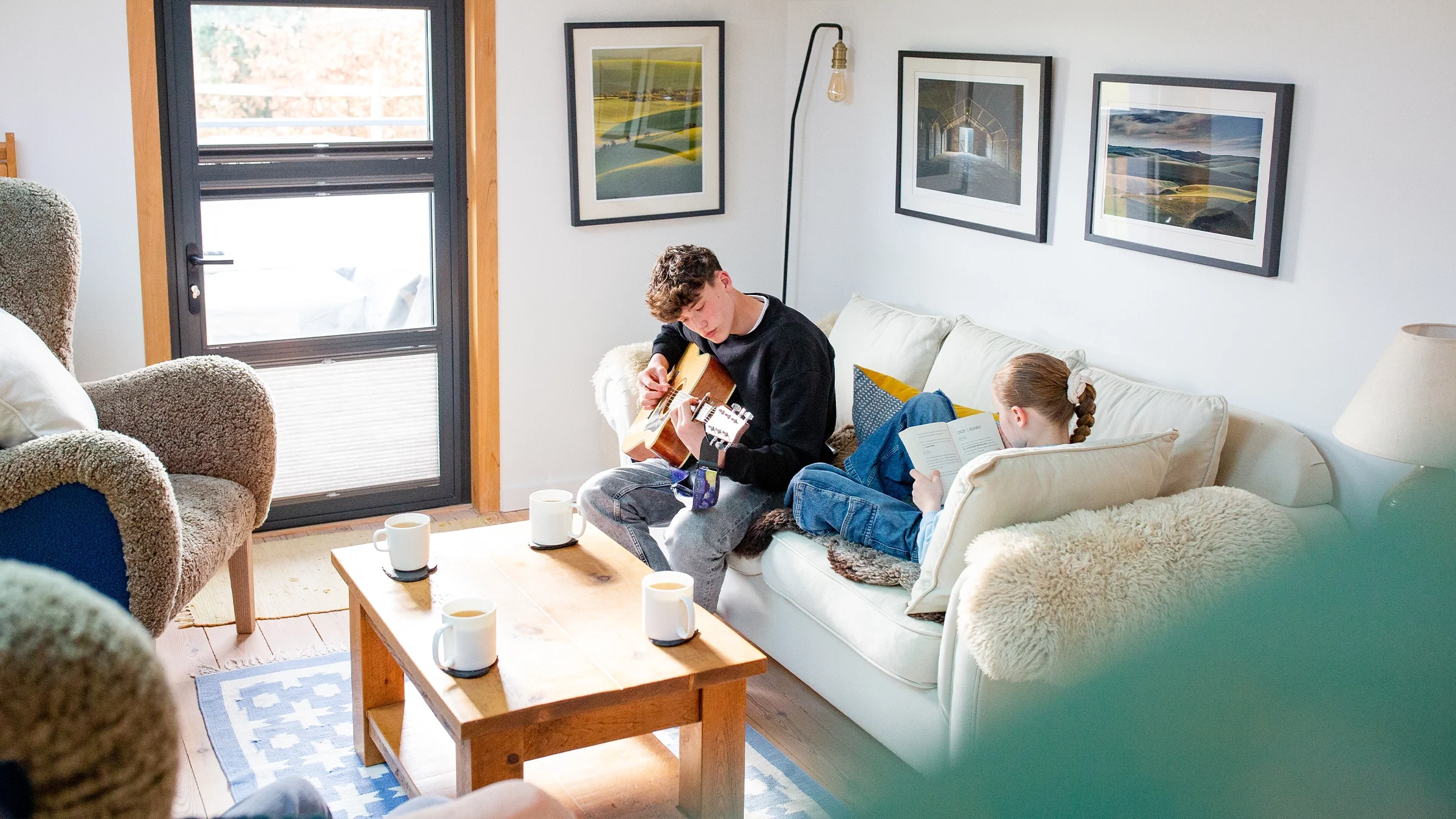 Two children sitting in a large, brown armchair reading a book together in a cozy, well-lit room with bookshelves and houseplants.