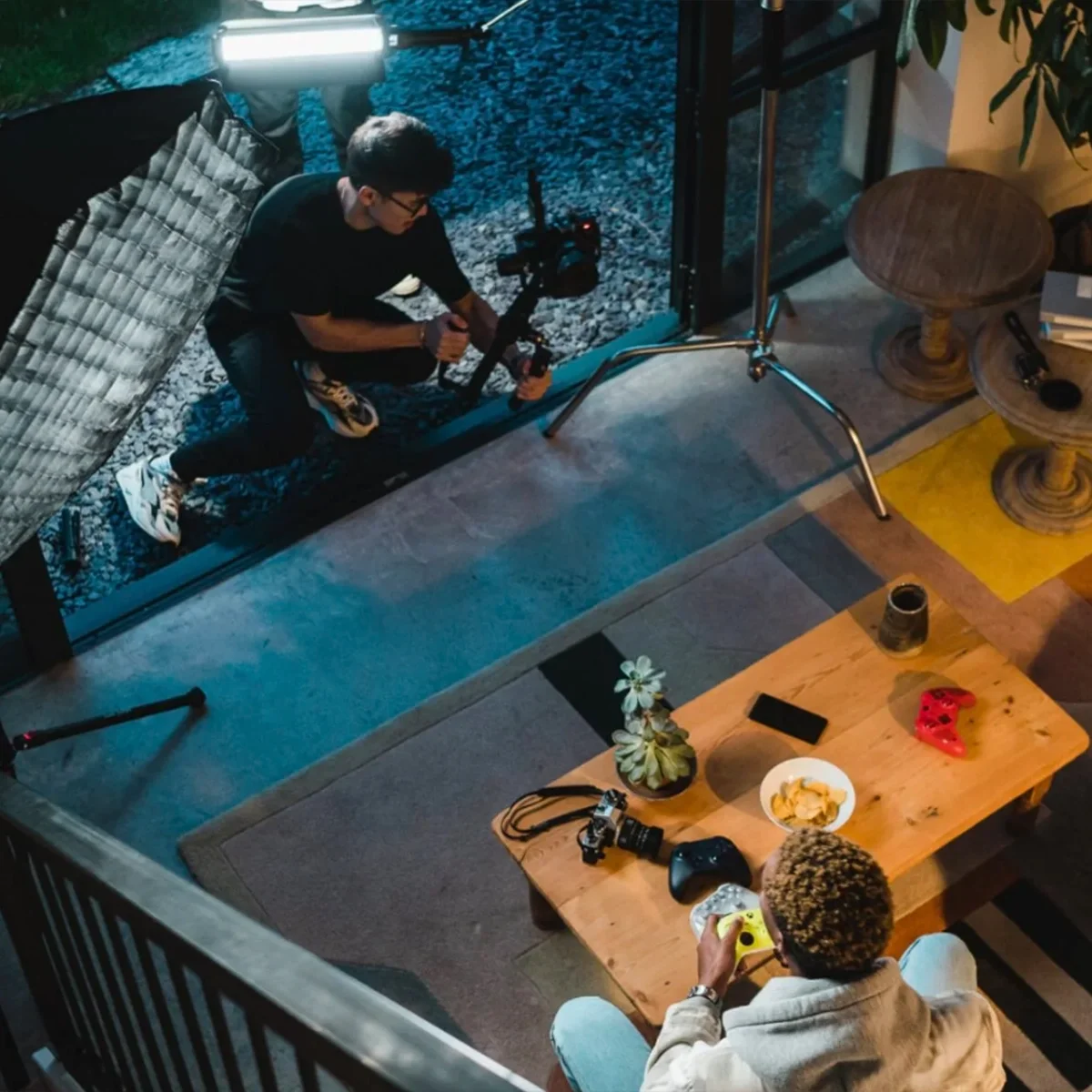 Indoor view of a living room with a person sitting on a couch playing video games with a controller, a bowl of snacks, camera, and gadgets on the coffee table. A videographer is filming from the outside through glass doors with lighting equipment set up.