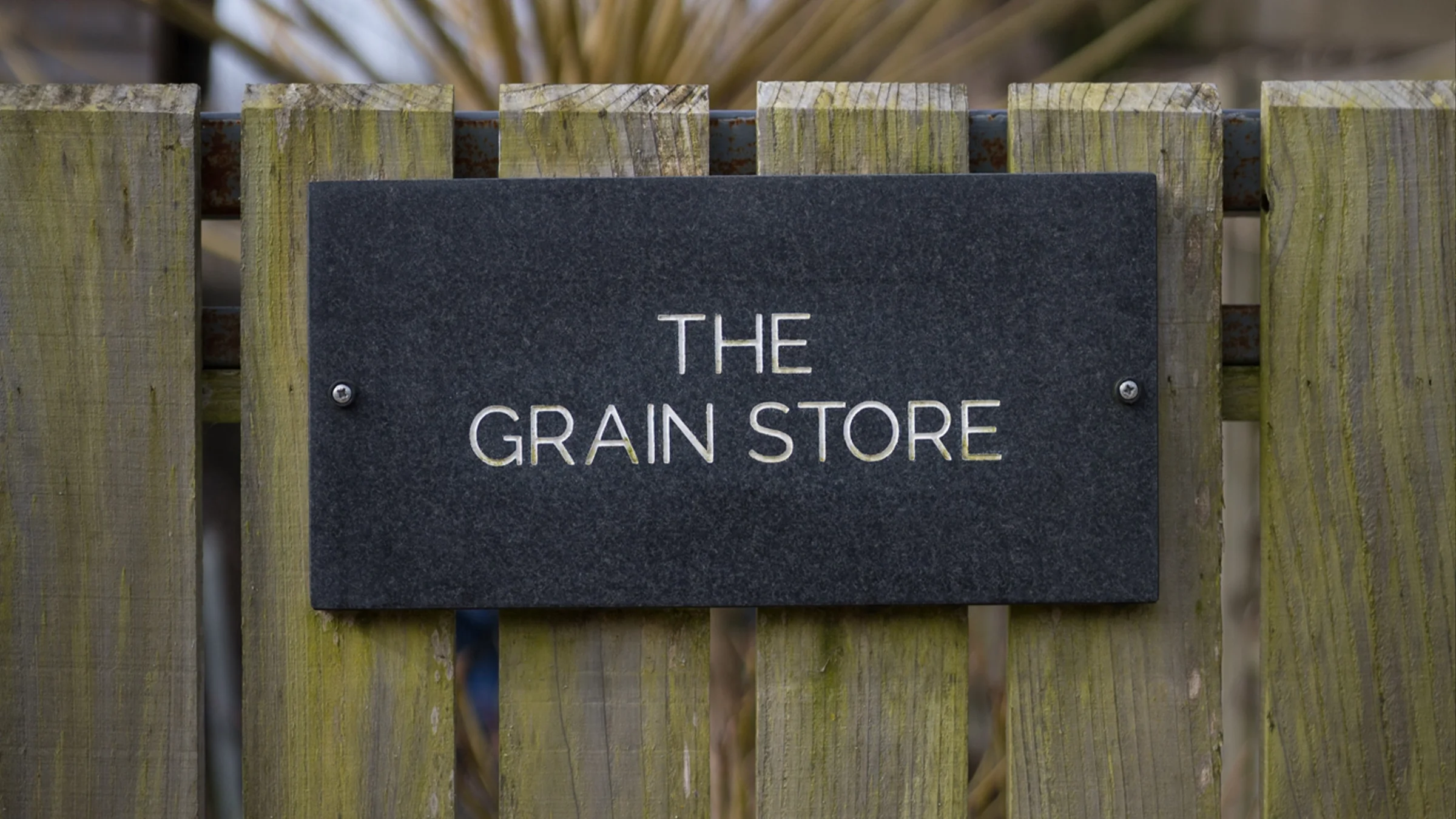 A black sign with white text reading 'The Grain Store' attached to a weathered wooden fence.