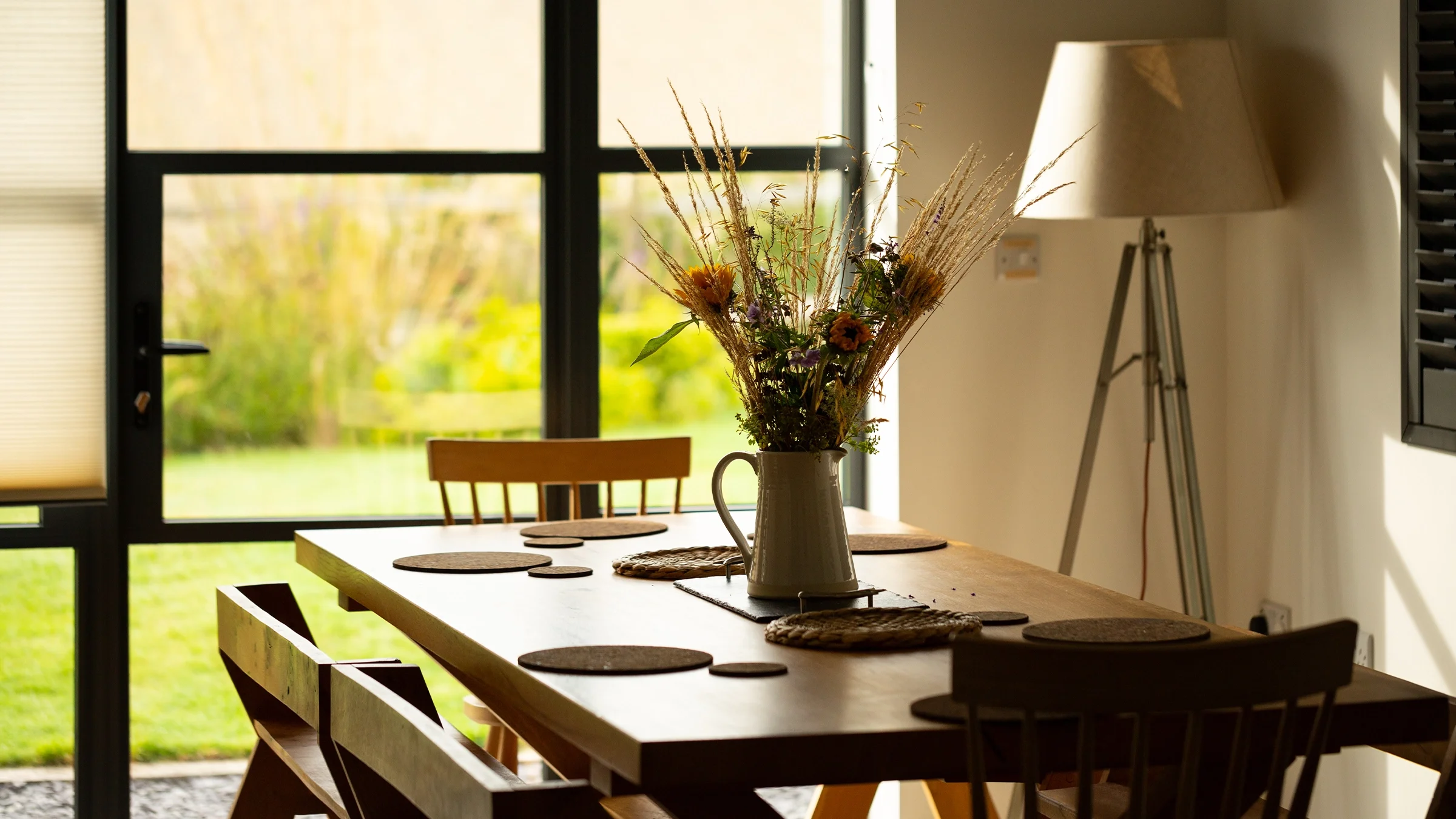 Modern living room and dining area with large windows, overlooking a garden. Two people sit at a wooden dining table having drinks. A person is relaxing on a sofa. Fruit bowl on the kitchen counter, TV in the background.