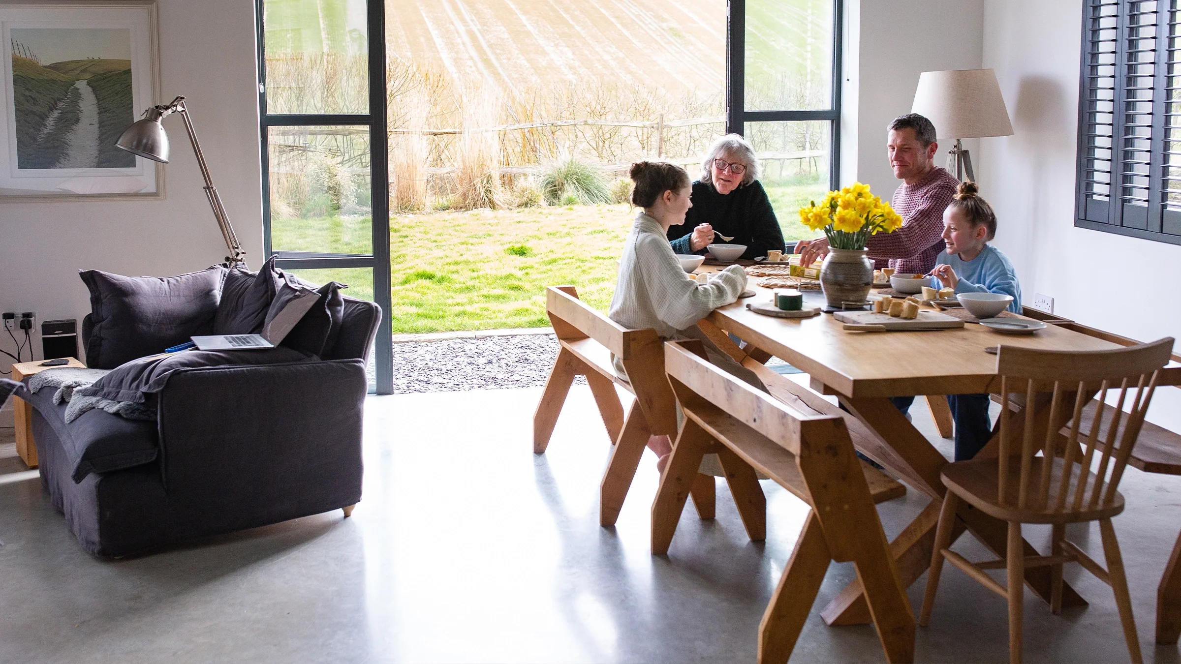 Family eating breakfast in a modern dining room with large windows overlooking a field, featuring wooden furniture, a bowl of fruit, and decorative plants.