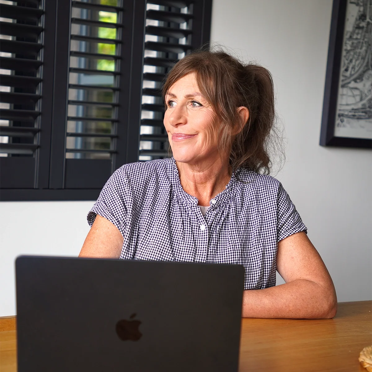 A woman with brown hair in a ponytail, wearing a checkered blouse, sitting at a wooden table with a closed laptop in front of her, smiling while looking to her left, in a room with black window shutters and a framed picture on the wall.