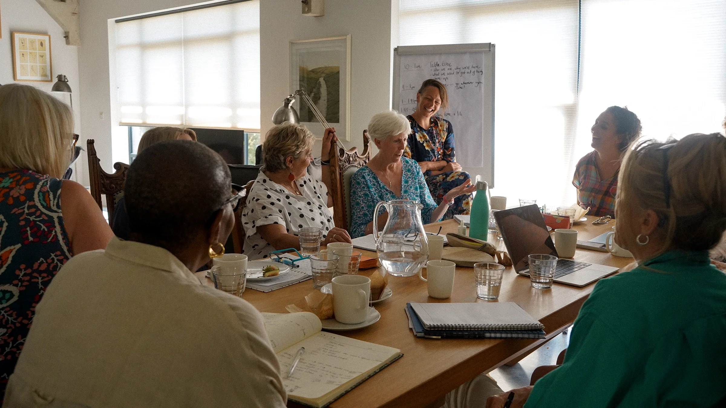 A group of women sitting around a conference table, smiling and engaging in conversation during a meeting or workshop, with a woman standing near a whiteboard.