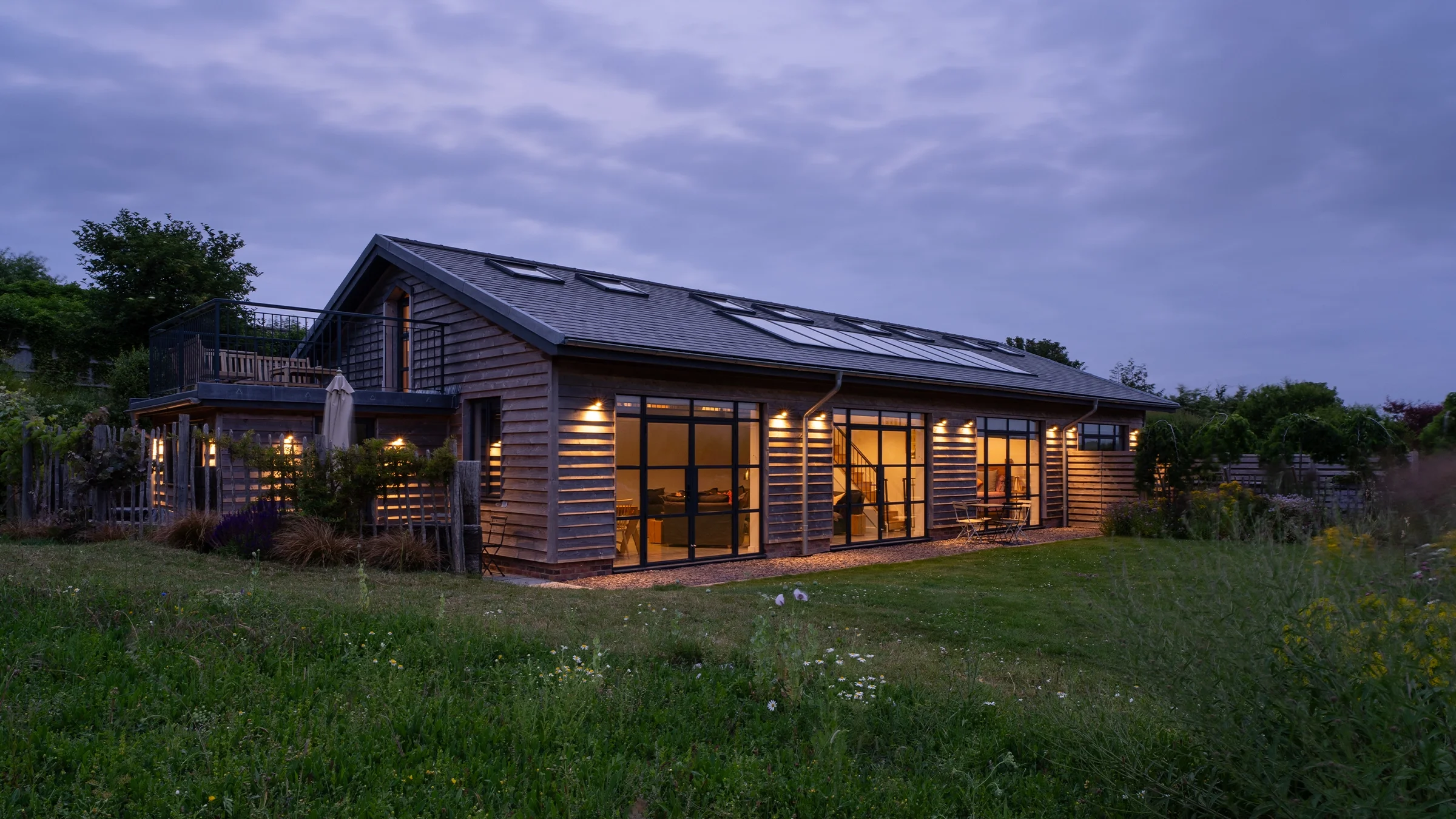Modern wooden house with solar panels, surrounded by a lush green lawn and wildflowers, featuring a balcony, deck chairs, and a person near a hot tub.