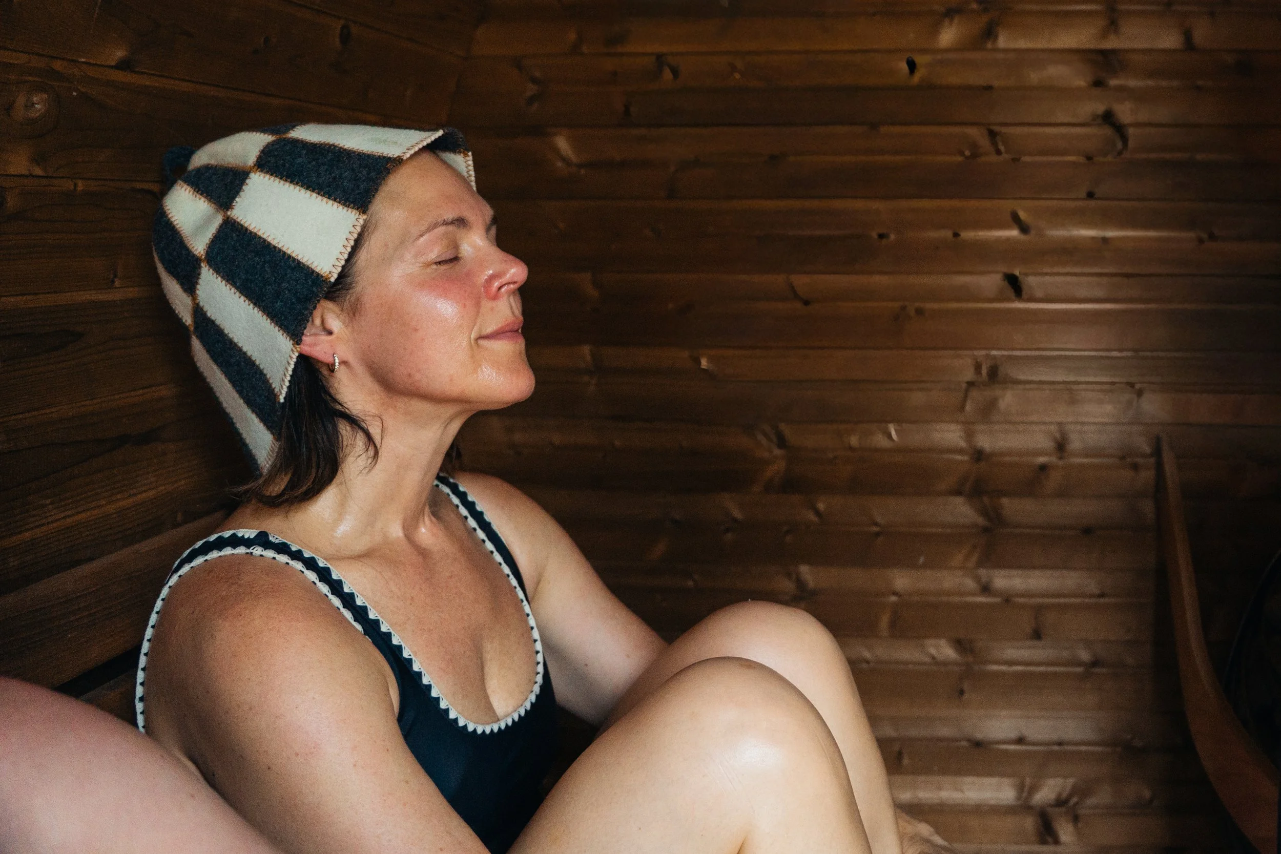 A woman with dark hair and earrings sitting with her eyes closed in The South Dows Sauna at The Grain Store Lewes, wearing a plaid towel on her head and a striped tank top.