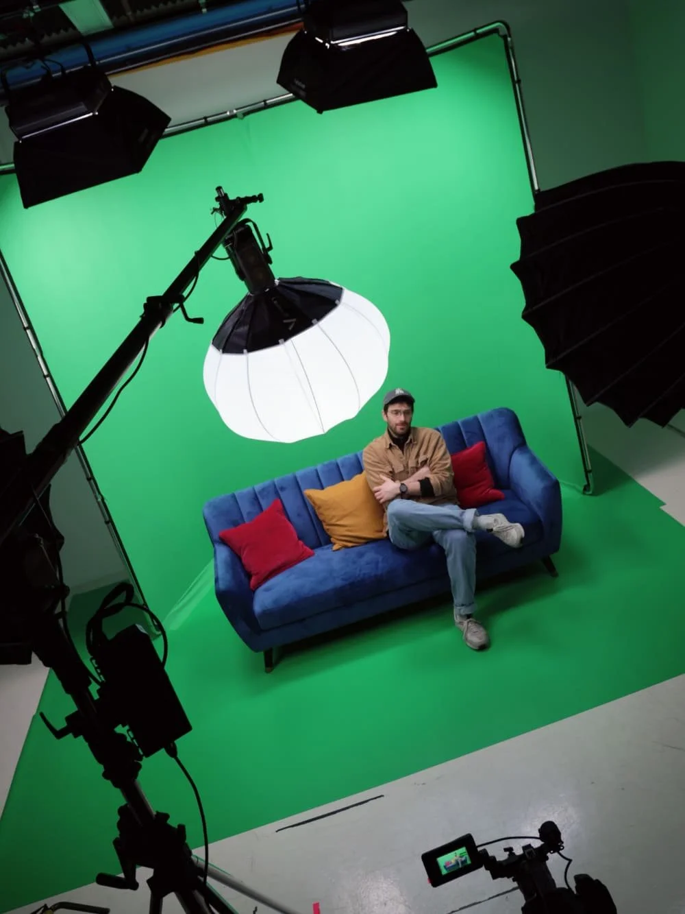 A man sits on a blue couch with colorful pillows in a Green Screen Studio, surrounded by lights and camera equipment.