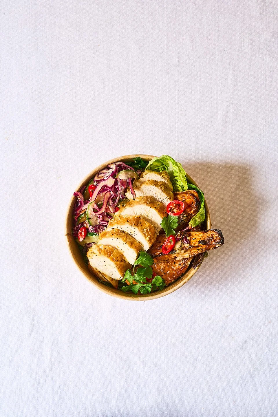 Bowl of grilled chicken slices, salad with purple cabbage and cucumber, lettuce, red chili slices, and herbs on a white tablecloth.