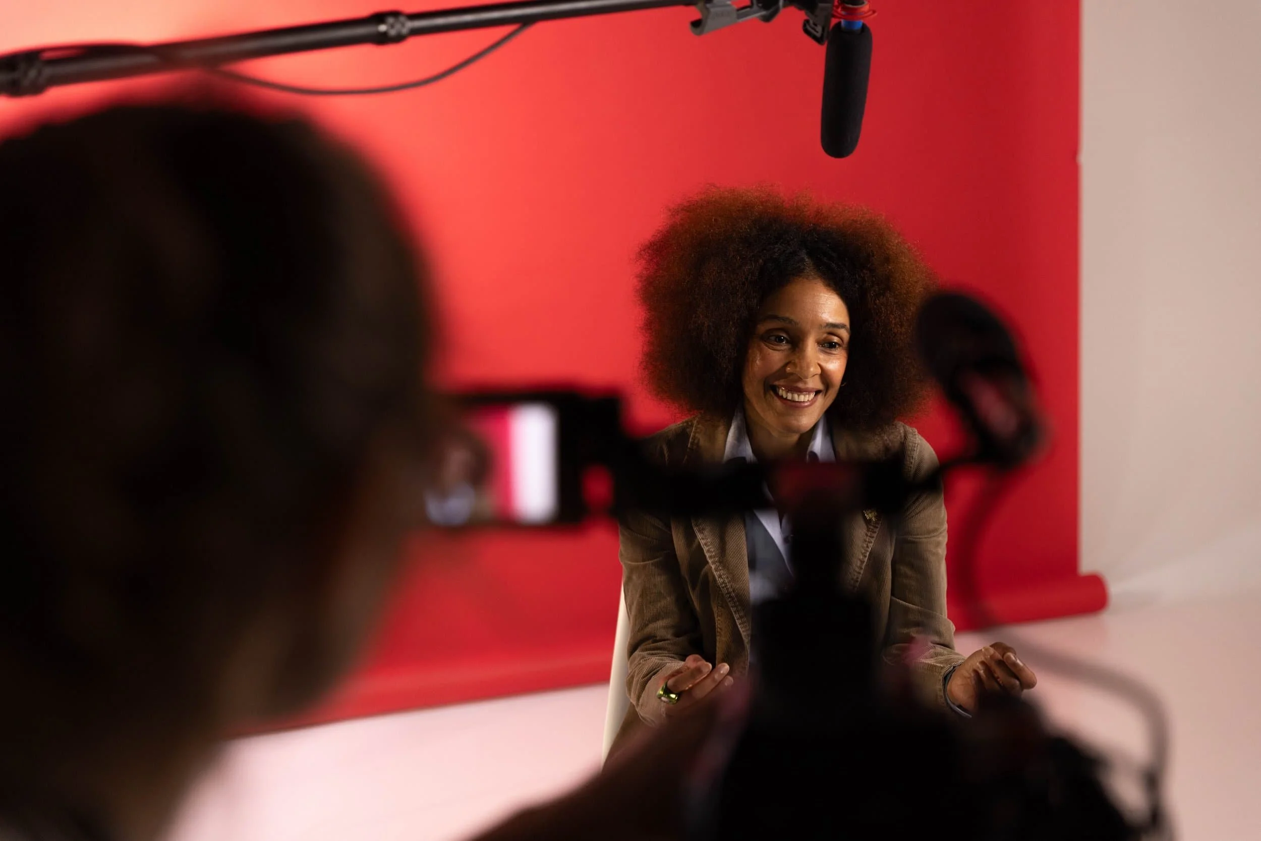 Filmmaker Rachel wang smiles in front of a red background. A microphone is above her head. In the foreground there is a blurred video camera