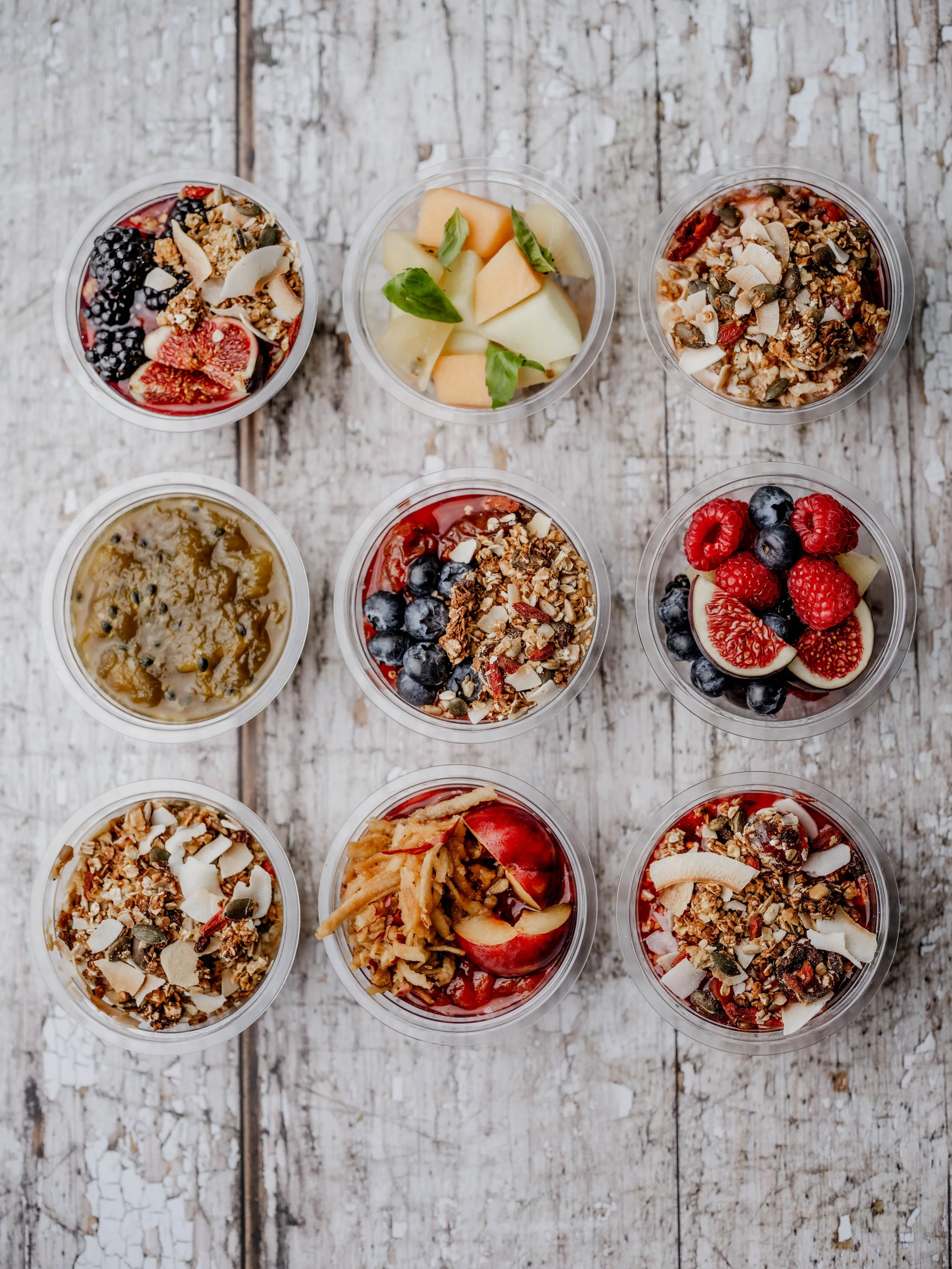 Nine small cups of yogurt parfaits topped with fruits, granola, coconut flakes, and other toppings arranged on a rustic wooden surface.