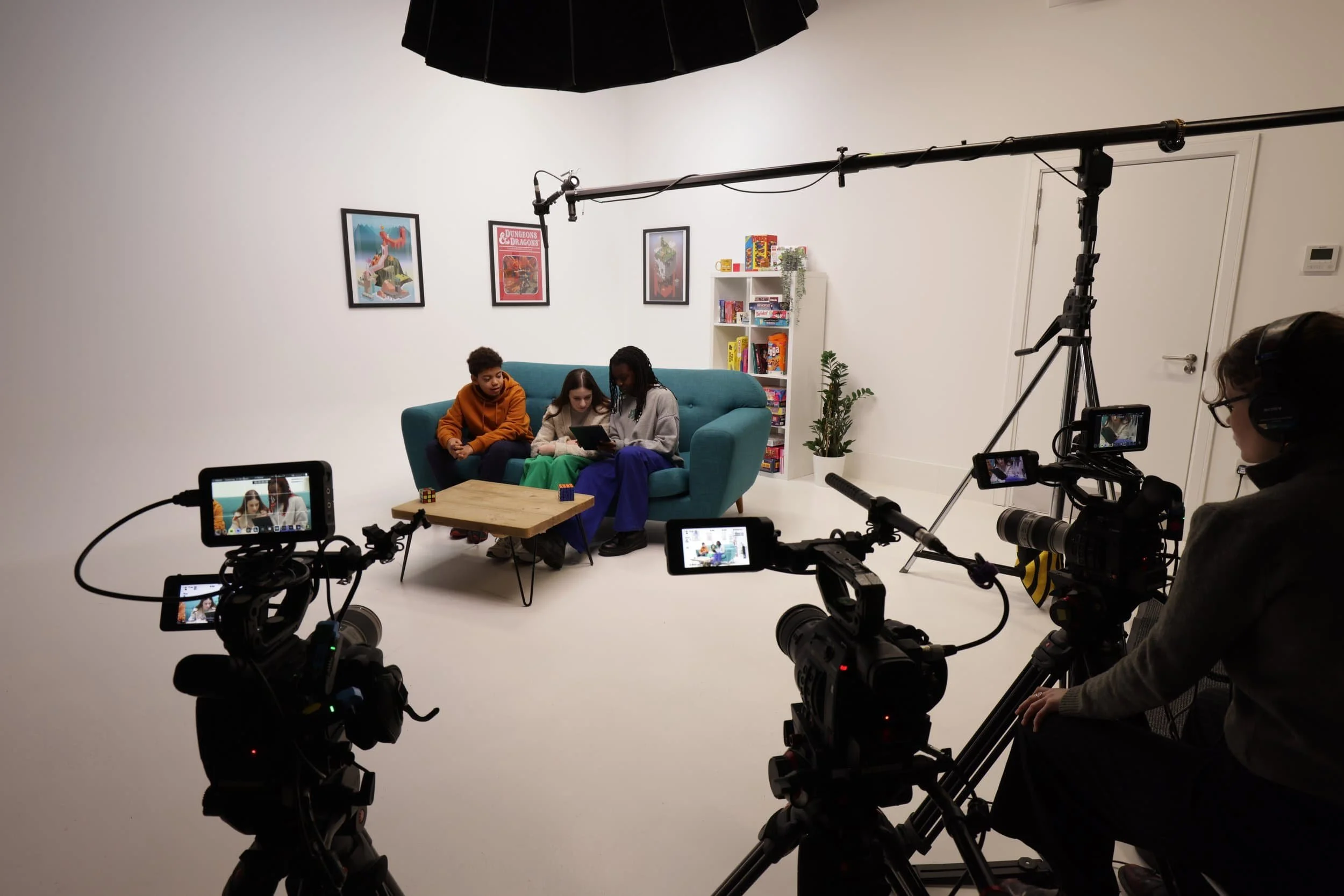 Three young people sit on a blue sofa. Three cameras film them as they look at an Ipad. There is a microphone above their heads.