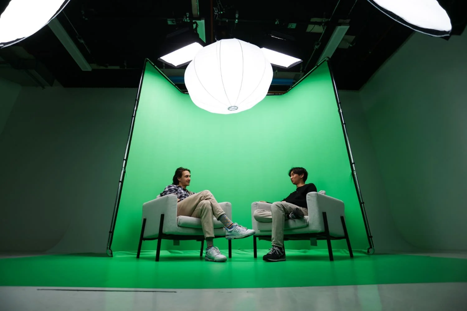 Two people sit in a Green Screen Studio, facing each other in chairs under bright overhead lights and ready for the shoot.