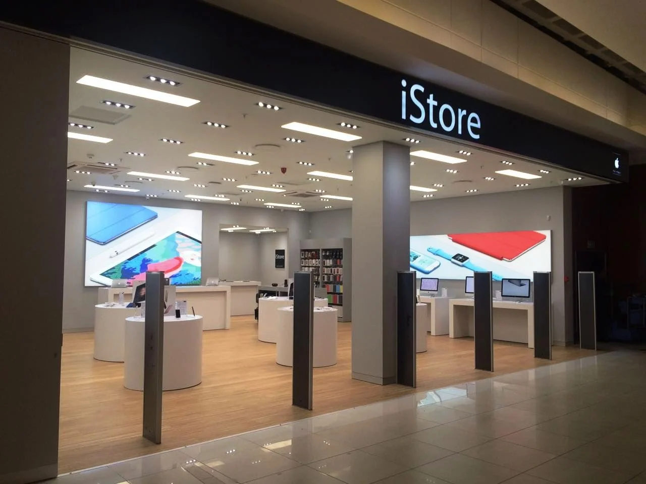Inside an Apple Store with display tables showcasing iPads and laptops, large digital screens on the walls, and a modern, well-lit design.