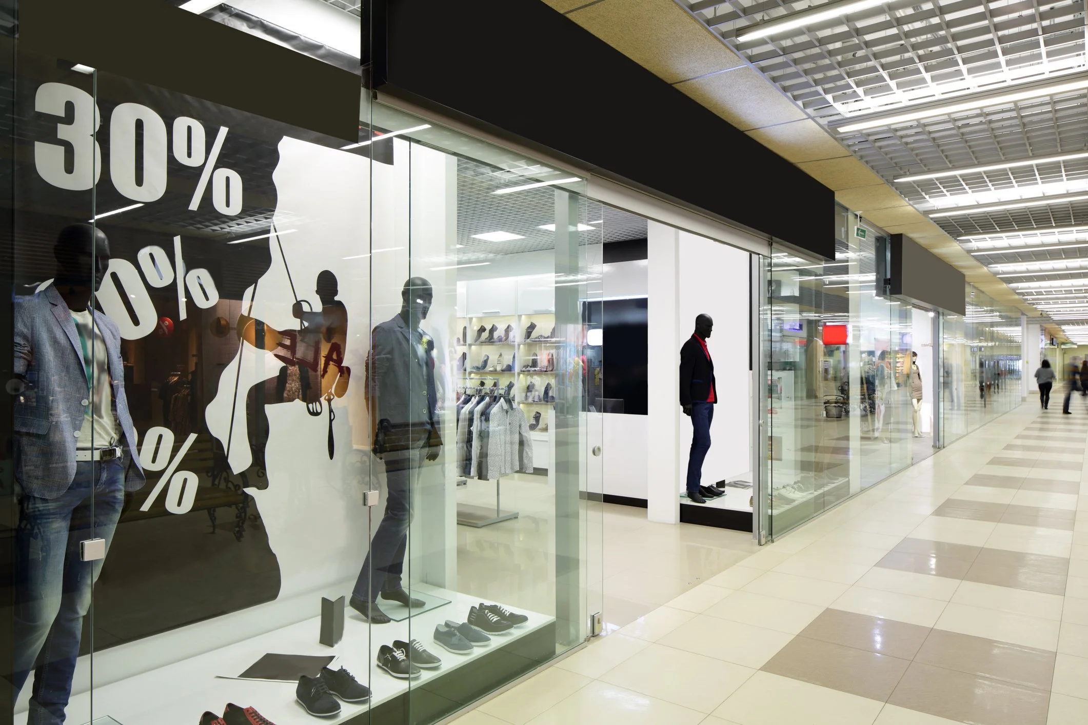 Mannequins in a shoe store window with discount signage, inside a shopping mall corridor.
