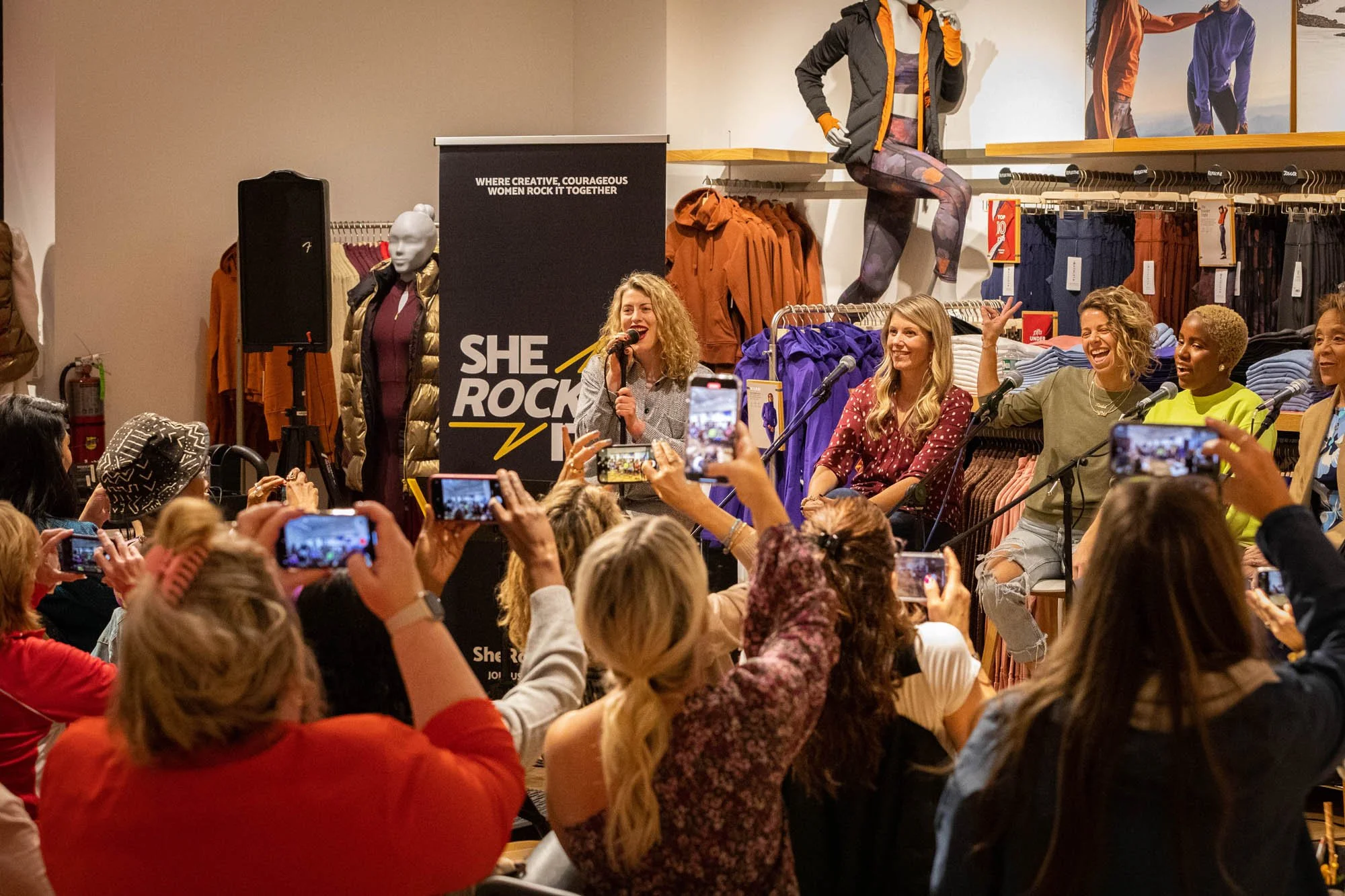 Women speaking and seated on a stage during a panel discussion at a retail store event, with an audience taking photos.