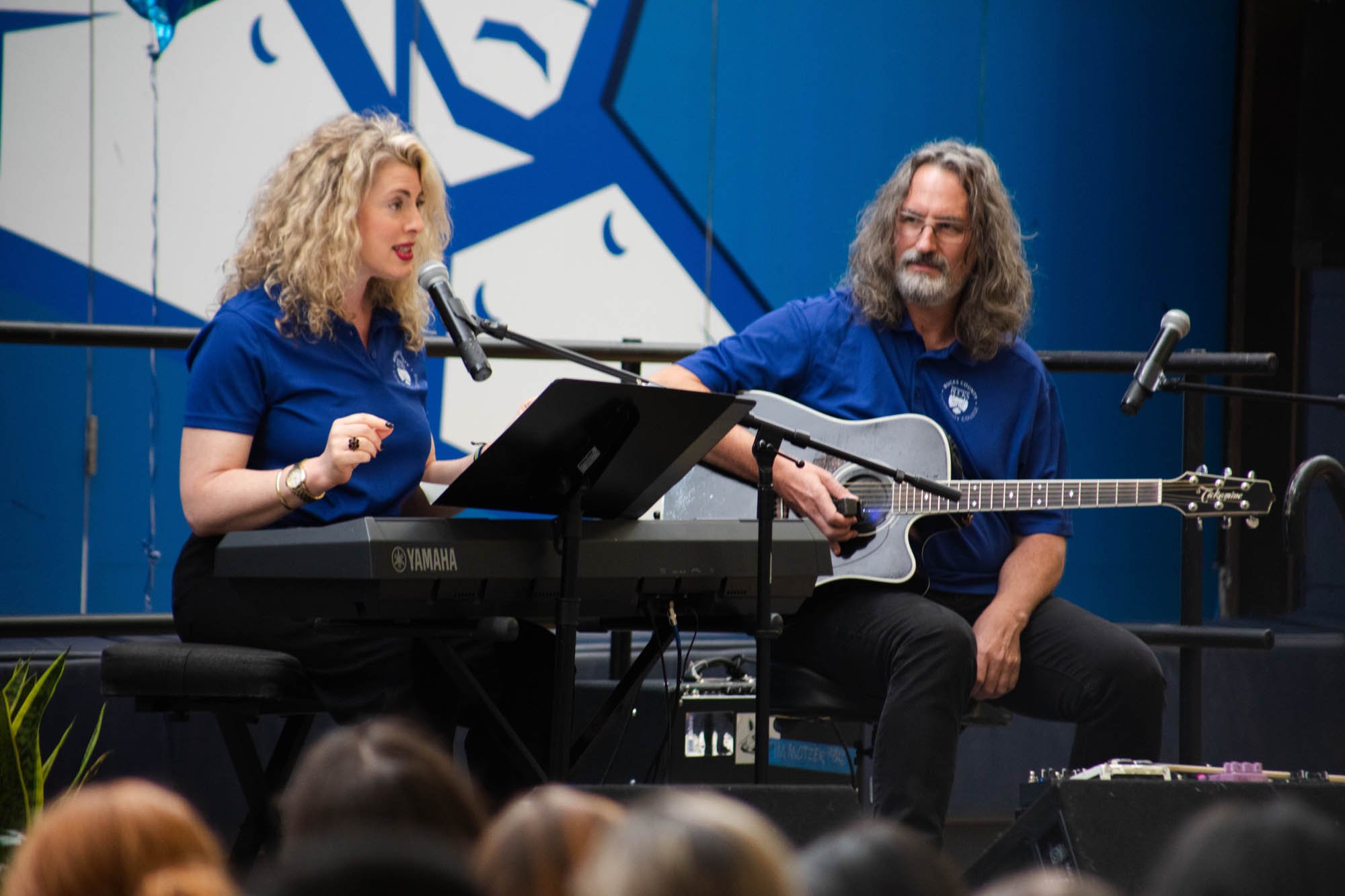 A woman singing into a microphone and playing a Yamaha keyboard, accompanied by a man playing an acoustic guitar, both wearing matching blue shirts.