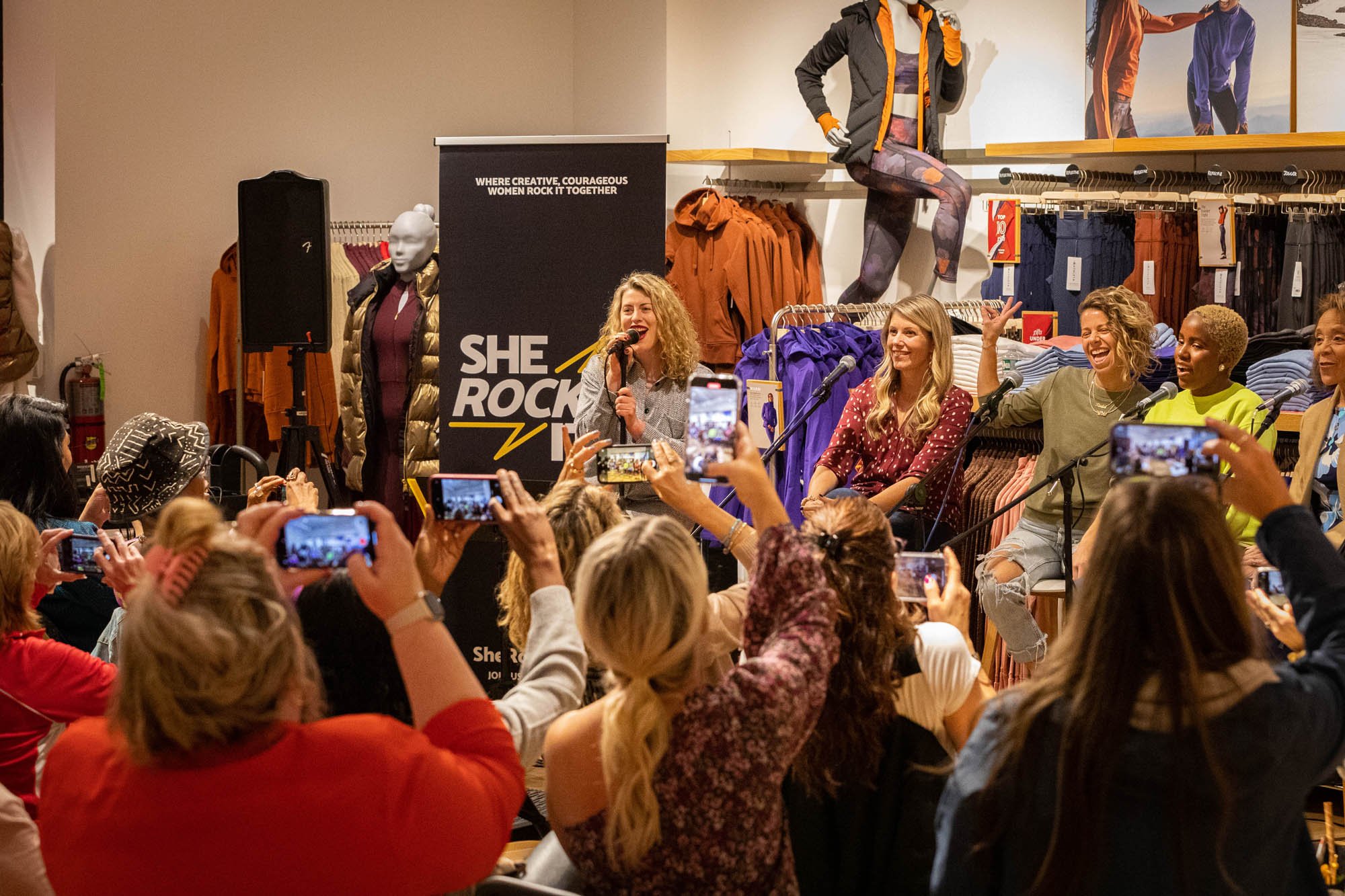 Women speaking and smiling at a panel discussion in a clothing store, with an audience taking photos.