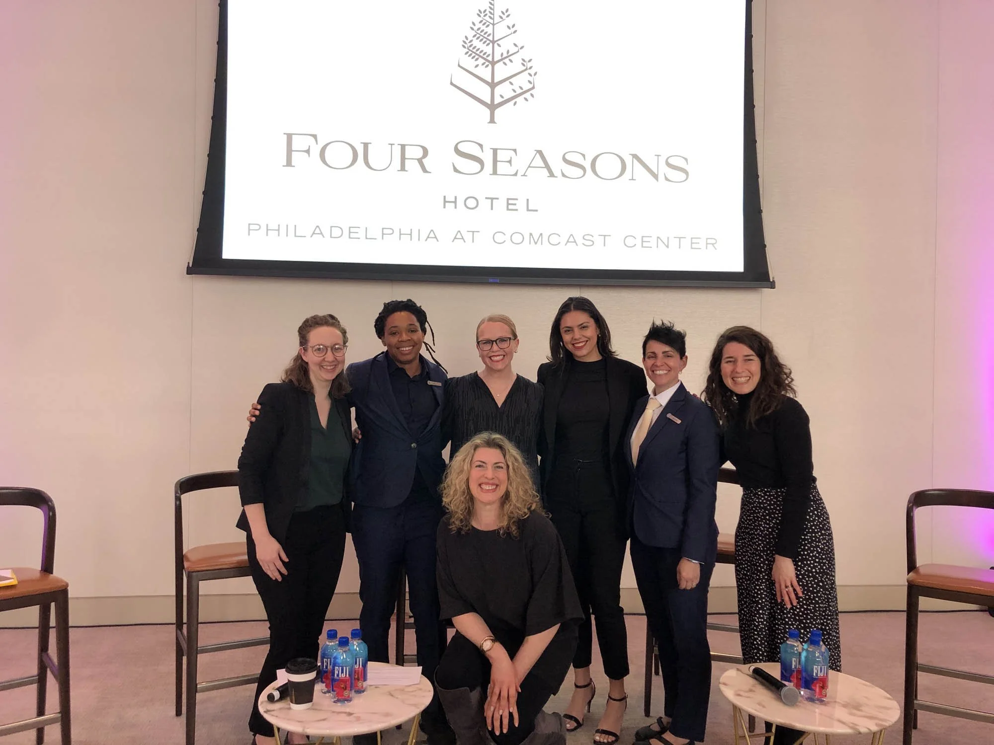 Group of seven women posing together at an event, with a large screen behind them displaying the logo for Four Seasons Hotel in Philadelphia at Comcast Center.