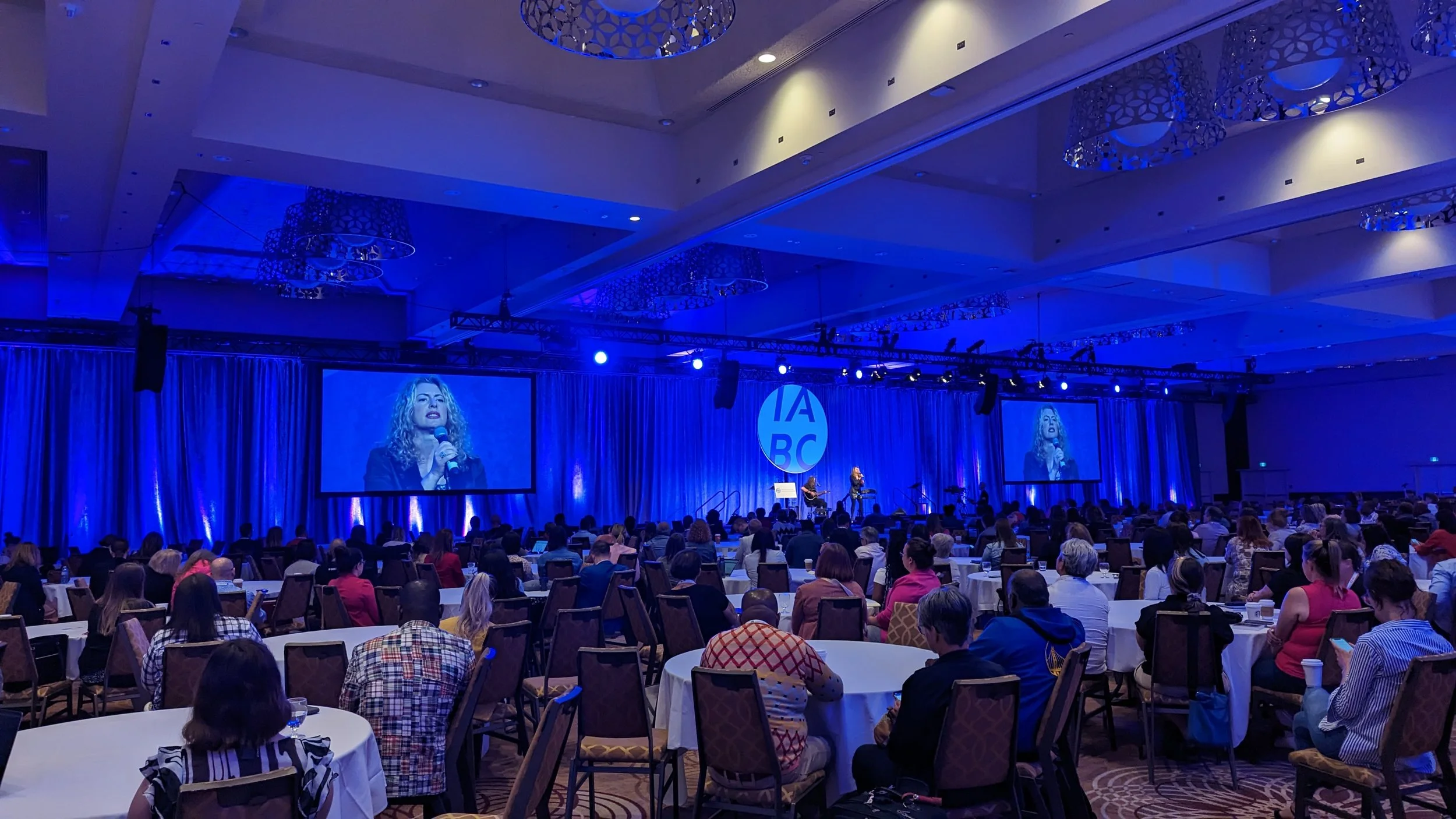 Large conference room filled with people seated at round tables watching a stage with a speaker on large screens, blue lighting, and decorative elements on the ceiling.