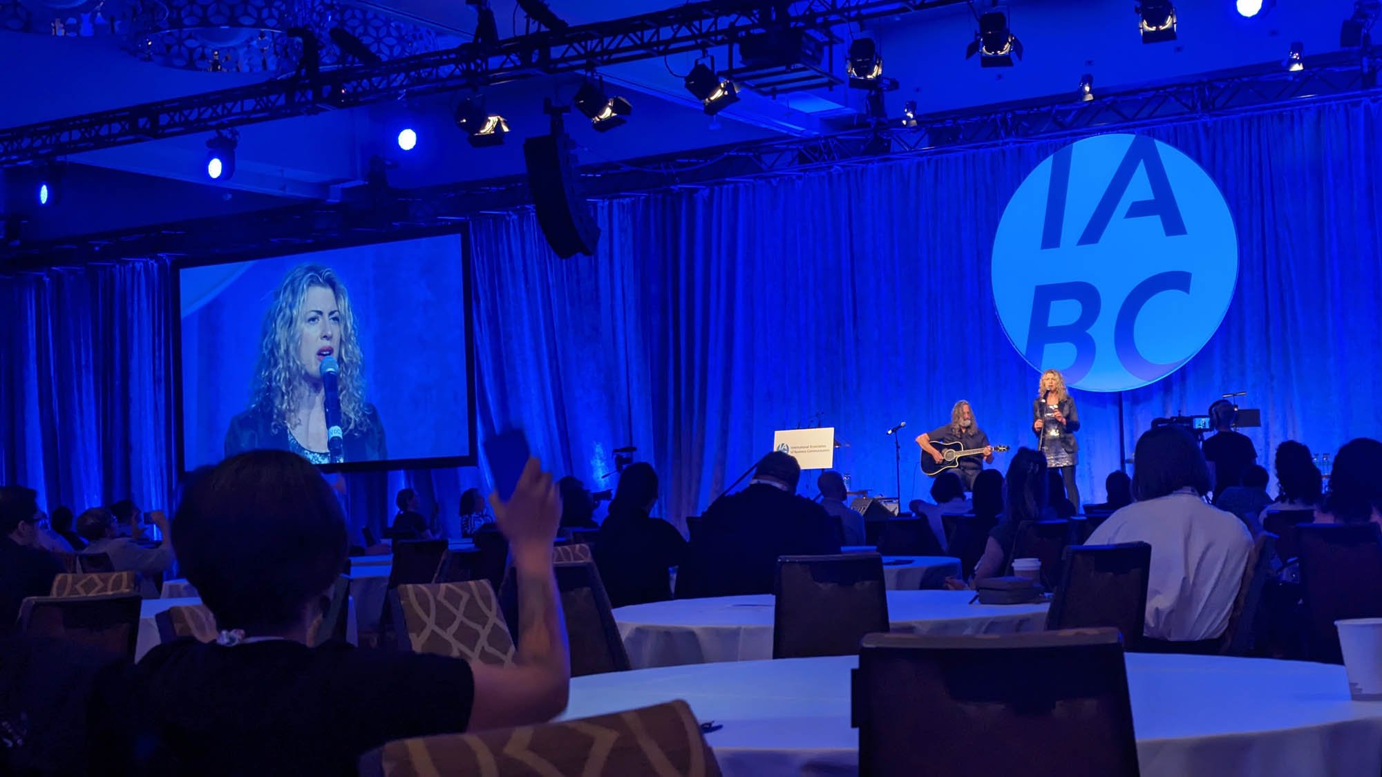 A woman singing into a microphone on stage with a guitarist. Large LED screen behind them displays her image. The stage has blue curtains with a circular logo that says 'IABC' in large letters. Audience members seated at round tables watch the perfor