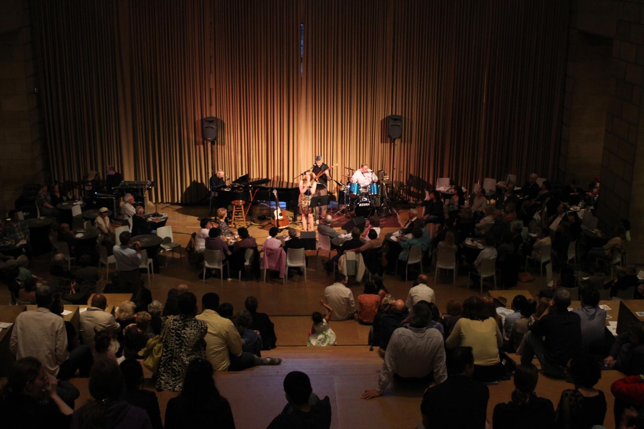 A live musical performance on stage with several musicians, including a singer, guitarist, bassist, and drummer, in front of a seated audience in a large concert hall or auditorium.