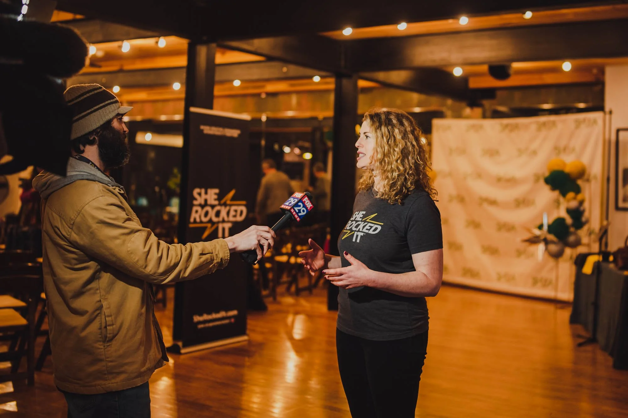 Woman being interviewed by a man holding a Fox 29 microphone indoors, with a black banner behind her reading 'She Rocked It' and some decor in the background.