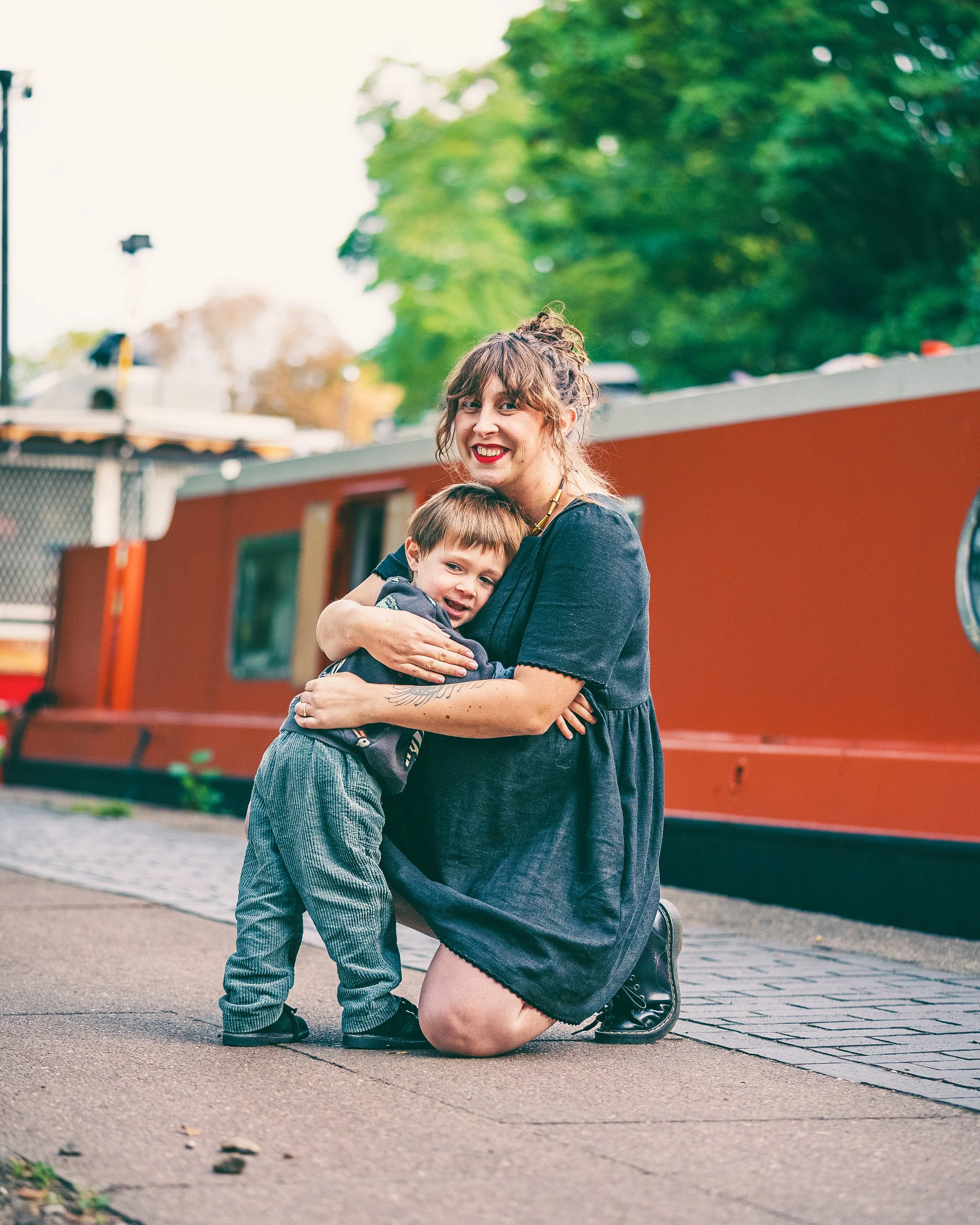 A woman kneeling on a sidewalk hugging a young boy in front of a red canal boat with trees and a fence in the background.