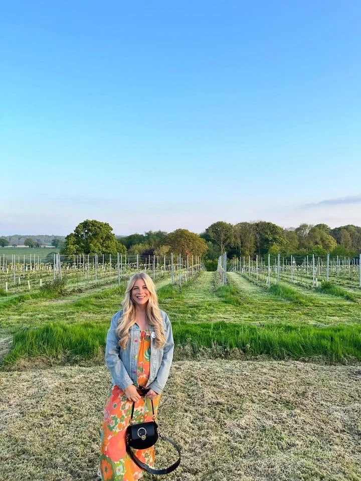 Young woman with long blonde hair smiling and holding a black purse, standing in front of a vineyard on a clear day.