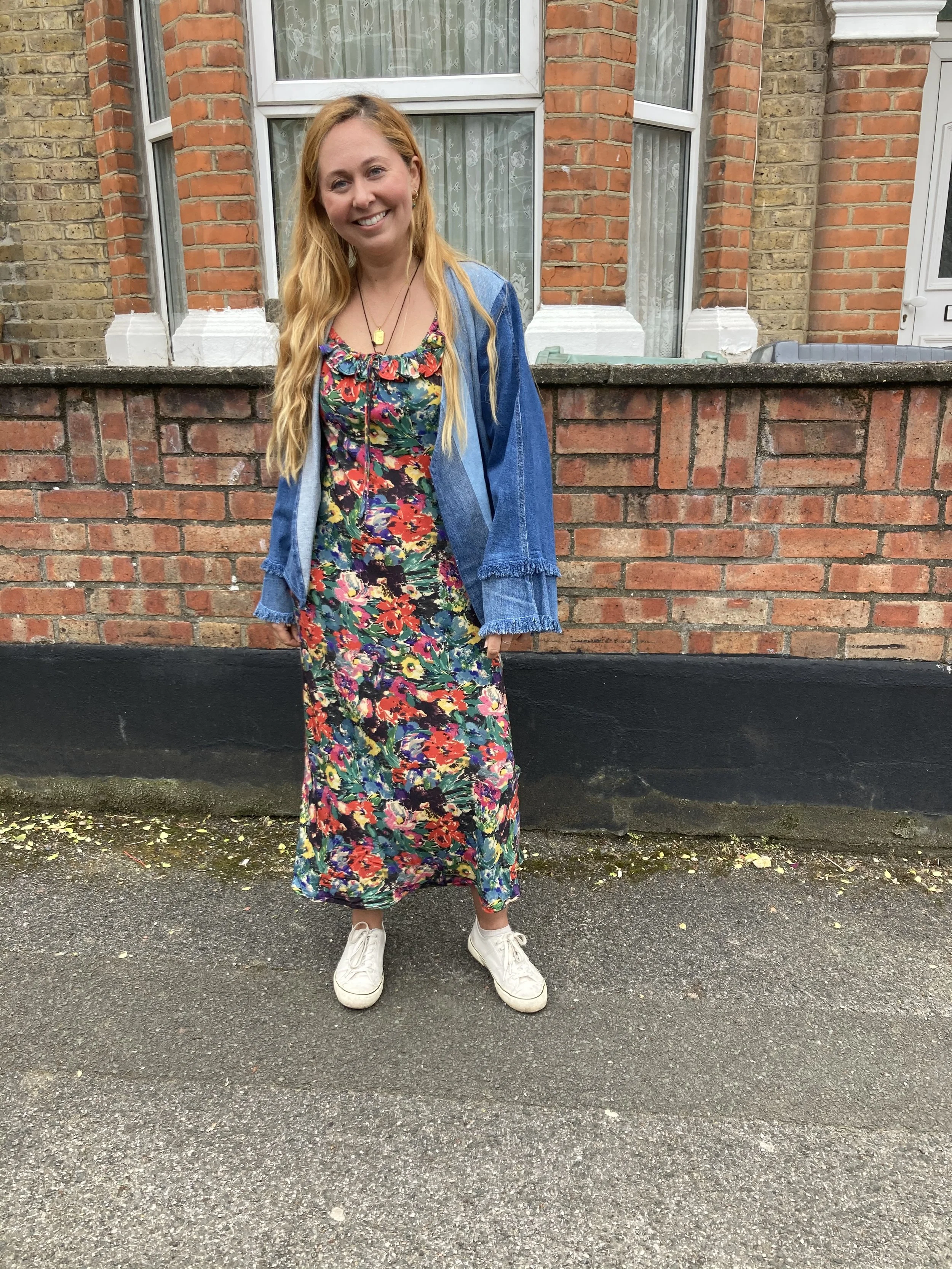A young woman with long red hair standing on a sidewalk in front of a brick wall, smiling at the camera, wearing a colorful floral dress, a denim jacket, and white sneakers.