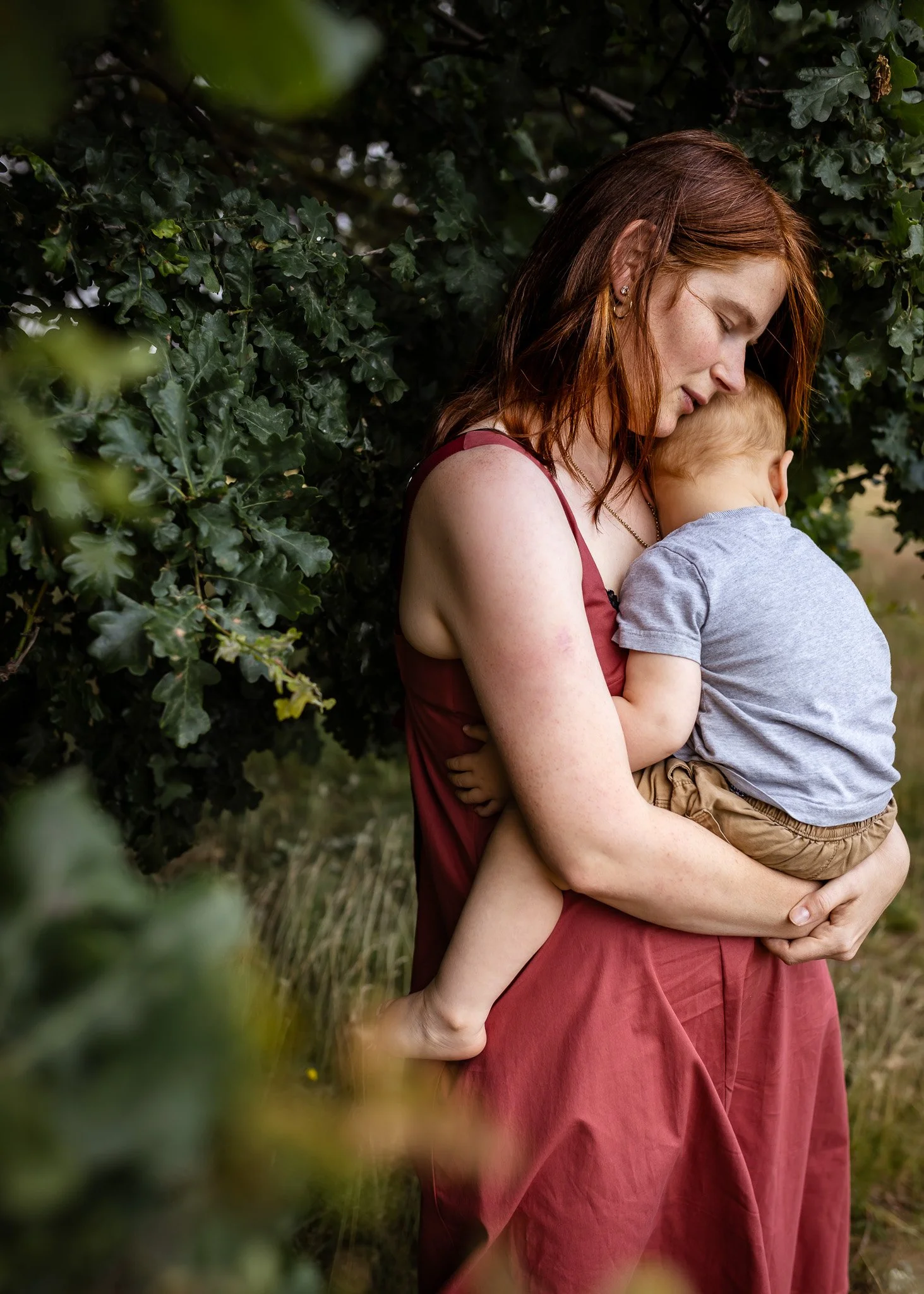 A woman with red hair holding a young child close to her face outdoors, surrounded by green foliage.