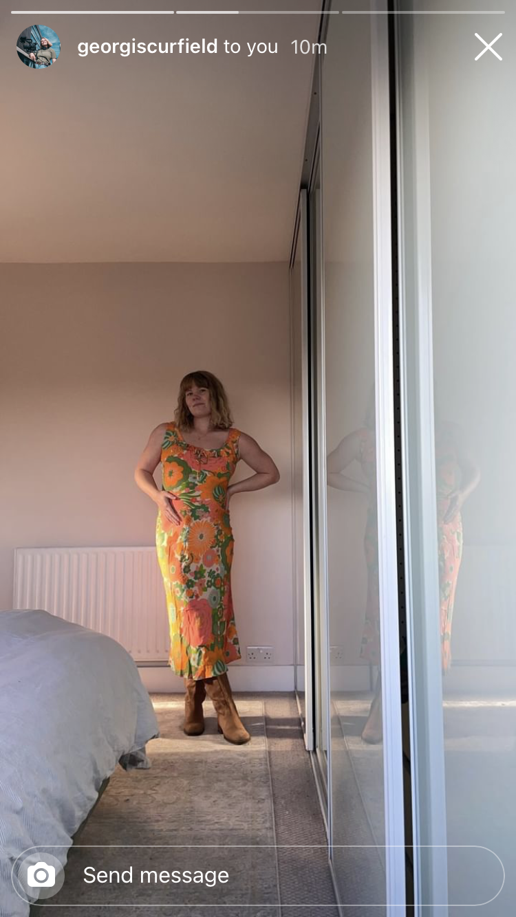 A woman standing in a bedroom, wearing a colorful floral dress and brown boots, posing with one hand on her hip and the other resting on her stomach, with a mirrored wardrobe reflecting her.