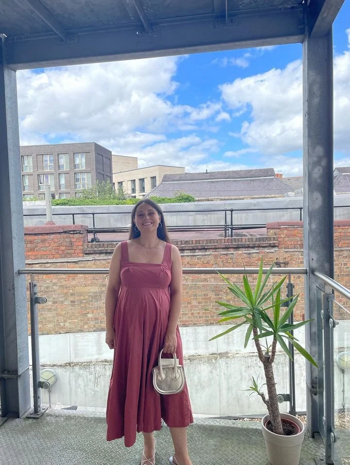 A woman in a pink dress holding a small beige handbag standing on a balcony with a potted plant, brick wall, and modern buildings in the background under a blue sky with clouds.