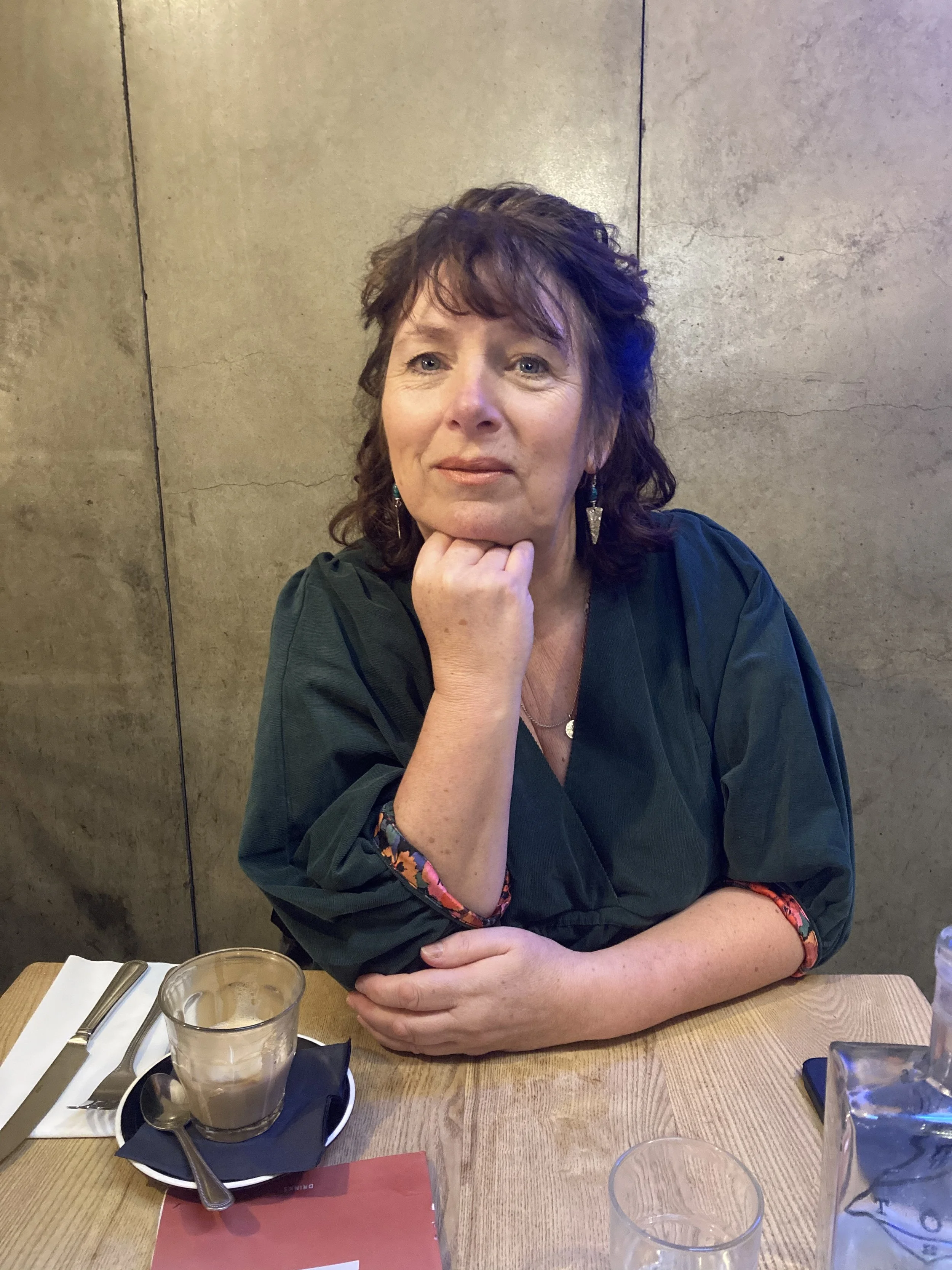 A woman with curly, dark brown hair and wearing a green top, sitting at a wooden table in a restaurant or cafe, resting her chin on her hand, with a thoughtful expression.