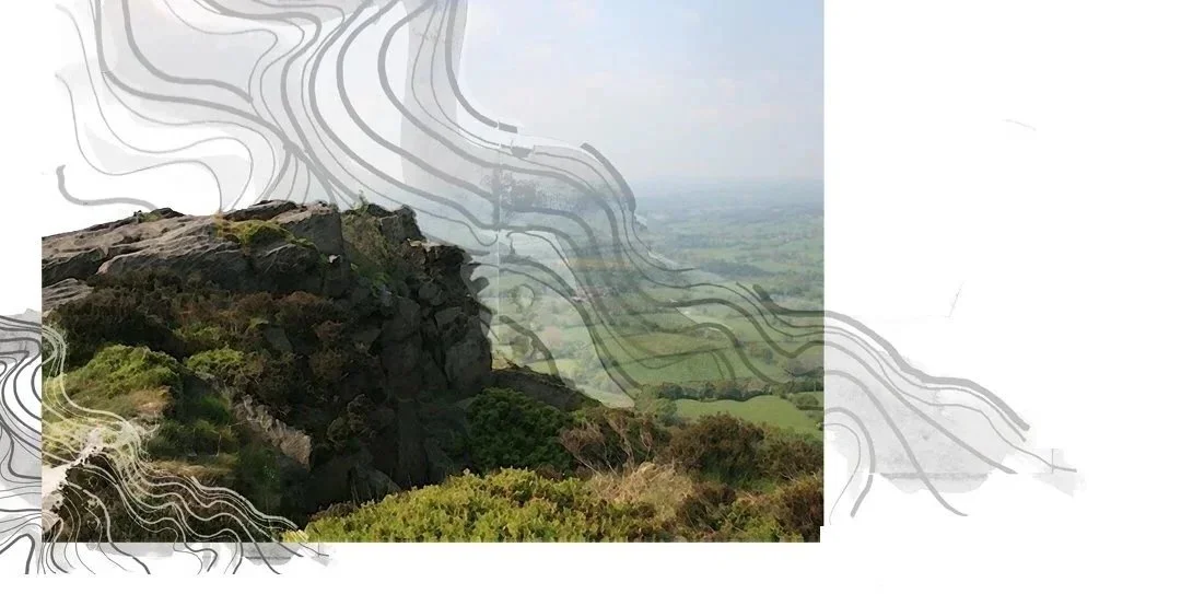 A rocky hilltop with green shrubs overlooking a green landscape with fields, under a cloudy sky.
