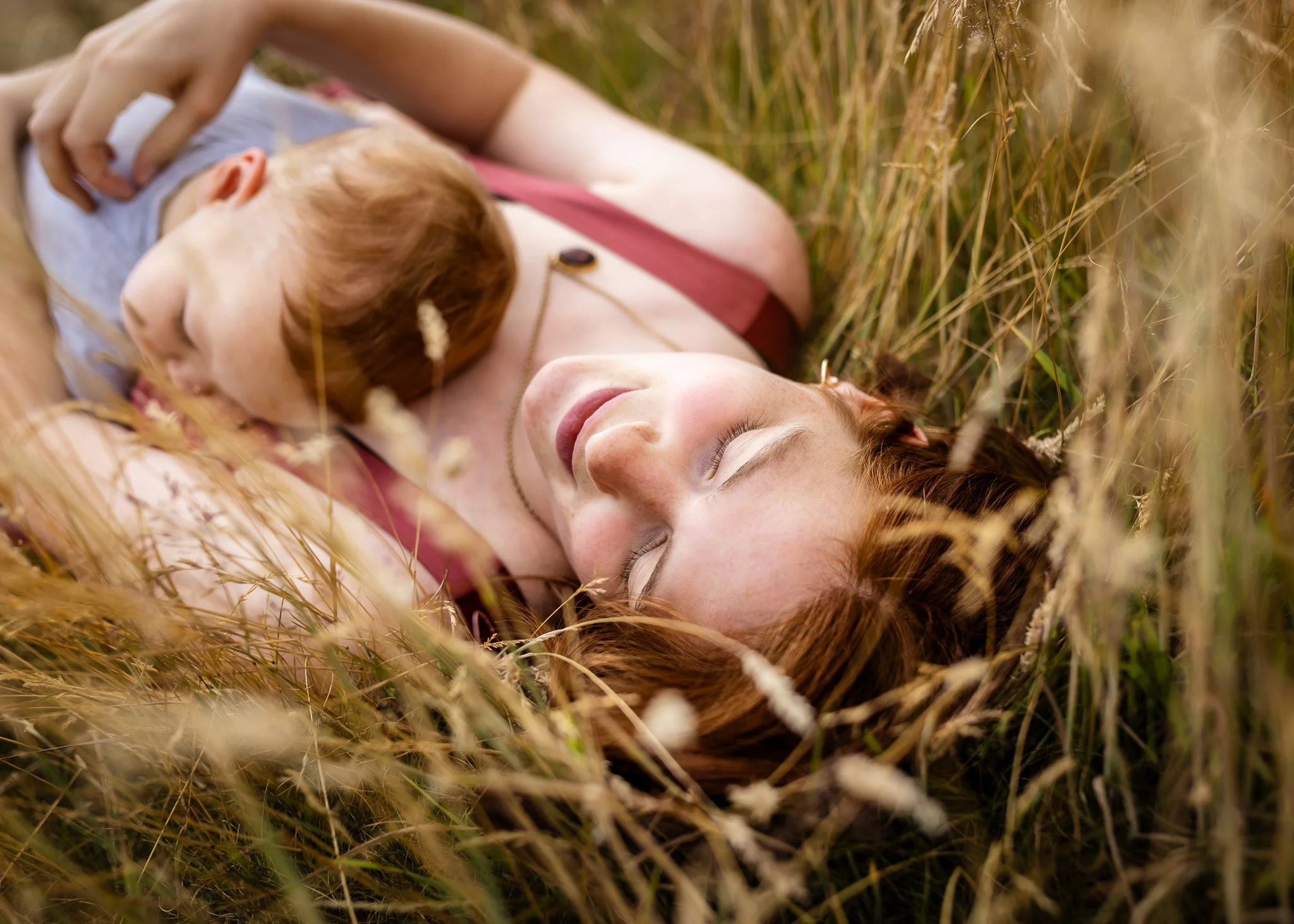 A young woman with closed eyes lying in a field of tall grass, holding a small child with red hair close to her chest.