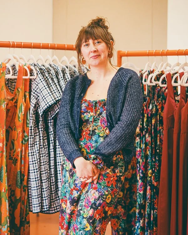 Woman standing in a clothing store surrounded by racks of colorful dresses, wearing a floral dress and a dark cardigan.