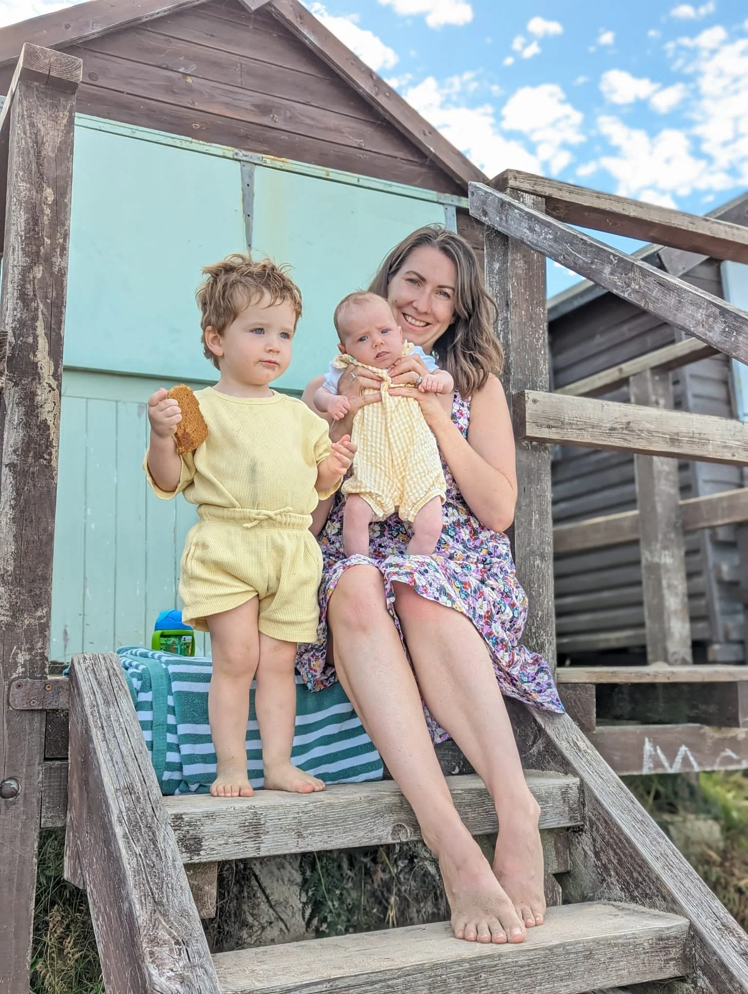 A woman with brown hair sitting on wooden stairs outside, holding a baby in yellow and white clothing, with a young child in yellow standing beside her holding a piece of bread. The background shows a wooden structure and a blue sky with white clouds.