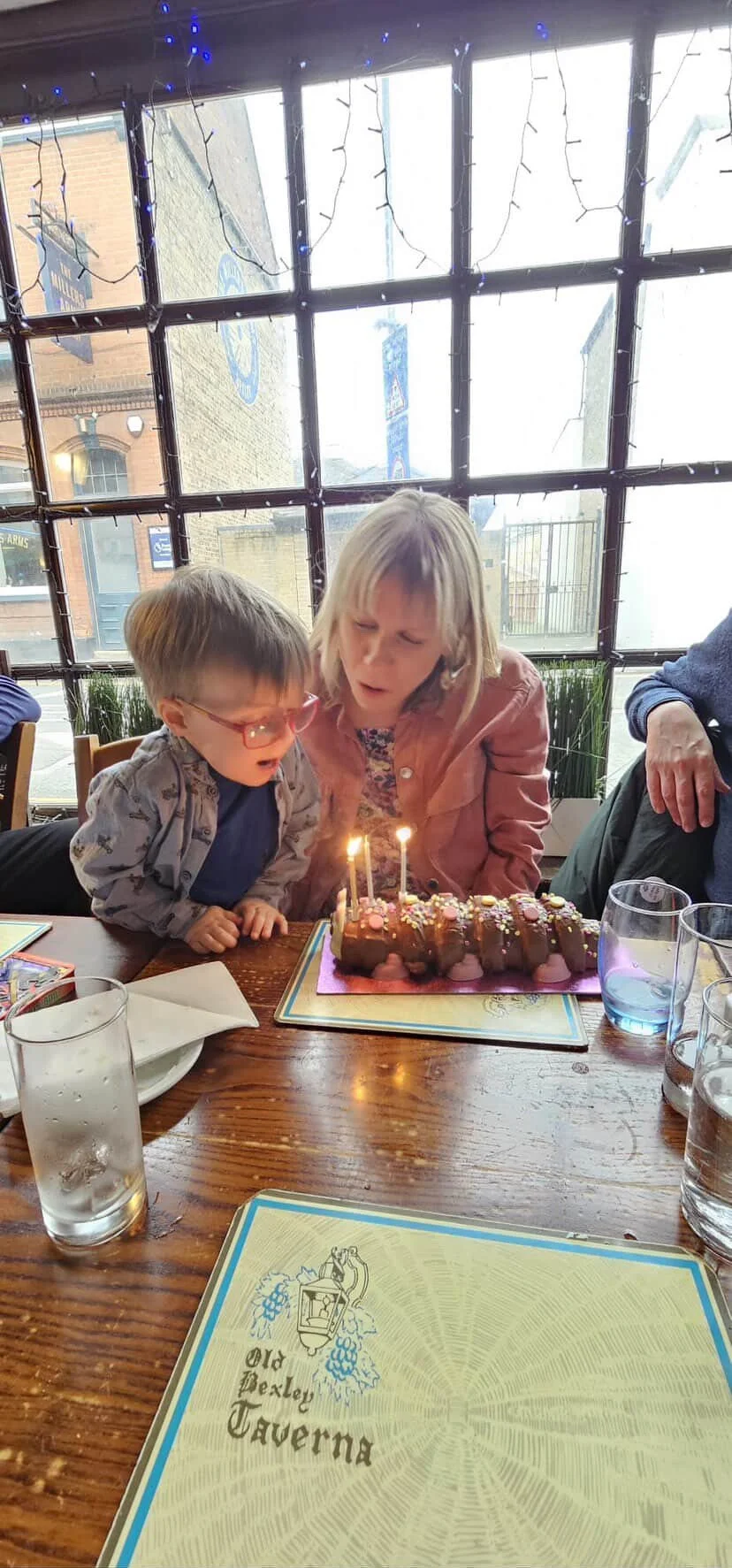 A woman and a young boy blowing out candles on a birthday cake inside a restaurant, with a window and holiday lights in the background.