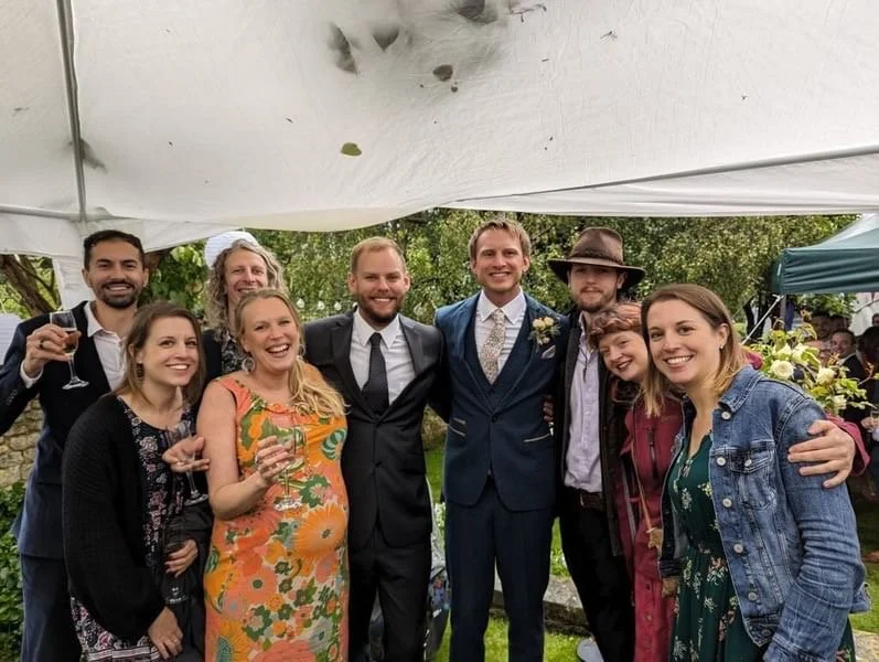 Group of eight people smiling and celebrating outdoors under a canopy, one man in a suit and tie, others in casual and semi-formal attire, holding drinks at a festive gathering.