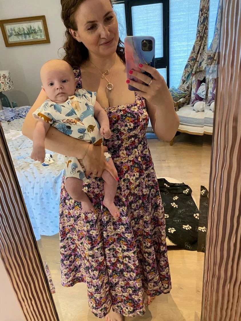 A woman with long brown hair in a floral dress holding a baby with a curious expression in her arms, taking a mirror selfie in a bedroom.