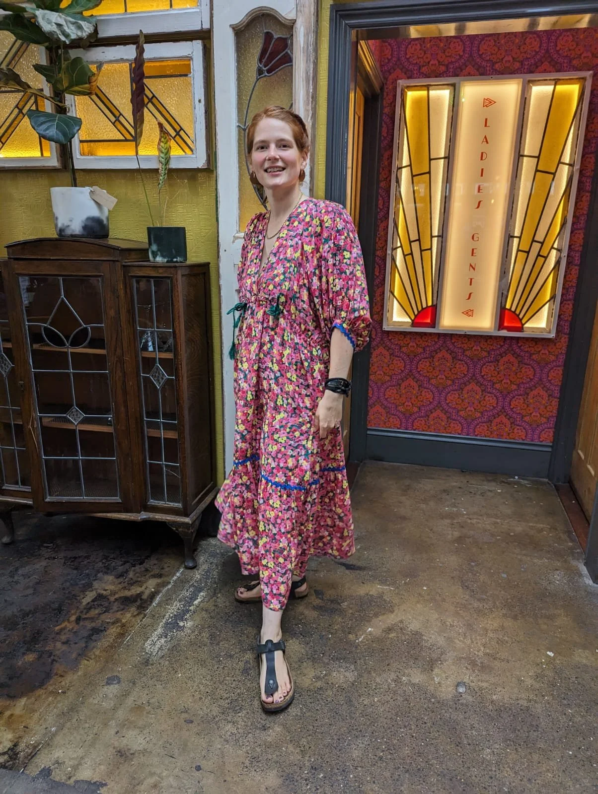 A woman in a colorful floral dress standing inside a vintage-style room near a door marked 'Ladies Gents' with ornate neon sign. She is smiling and wearing sandals.