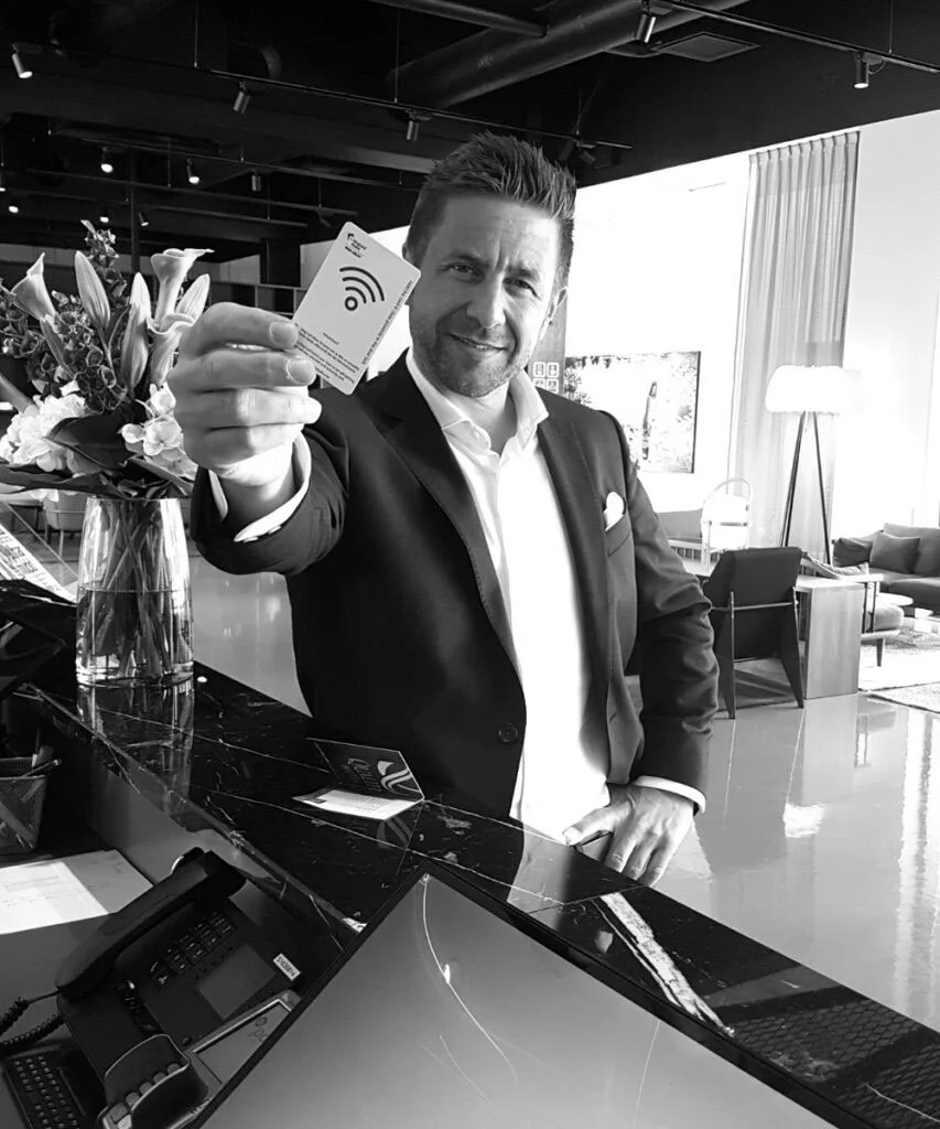 A man in a suit holding a card with a Wi-Fi symbol, standing at a reception desk in a modern hotel lobby.