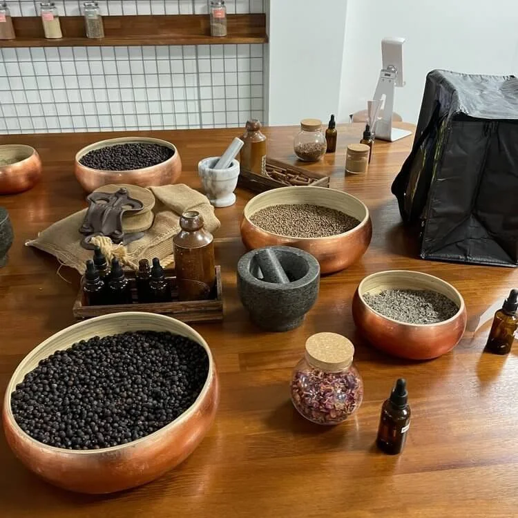 Baskets and jars of various herbs, spices, and seeds on a wooden table, with bottles and a black bag nearby.
