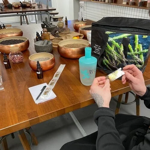 A person working with various herbs and spices on a wooden table, with copper bowls, small bottles, a bag of asparagus, a turquoise gin bottle, and a white tray with a pen and designed tape.